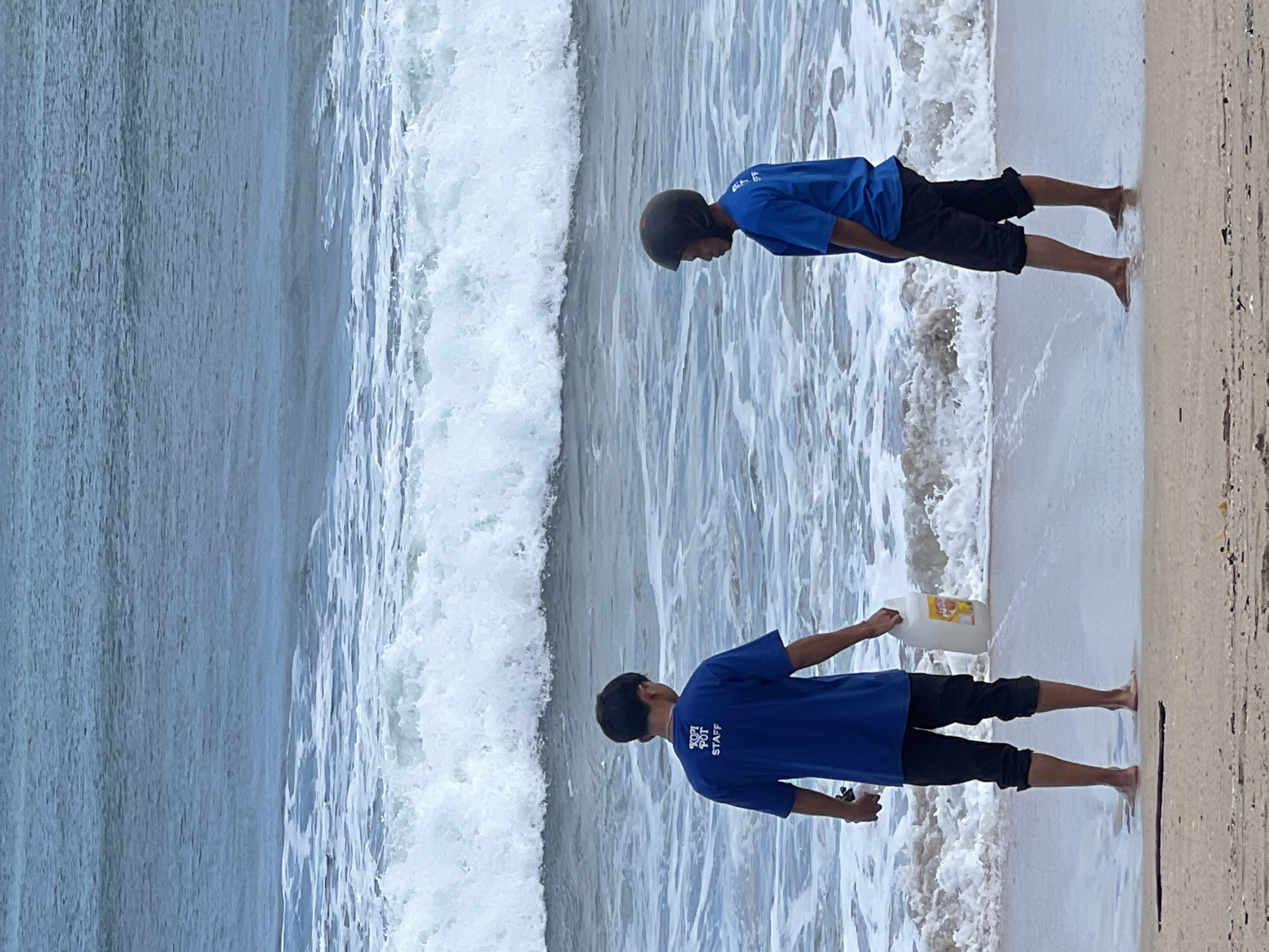 Two young males stand on the beach in Kuta, Indonesia, facing the ocean. The person on the left, seen from the back, wears a blue t-shirt with "KOPI POT STAFF" printed on the back and dark pants. They are holding a large white plastic jug with a yellow label in their right hand. The person on the right, also seen from the back, wears a blue t-shirt and dark shorts, and a dark helmet. Both individuals are standing in shallow water near the shoreline, with waves breaking behind them. The sand is light brown, and the ocean water is a mixture of blue and white foam. The overall scene depicts a casual moment by the sea, possibly involving work duties given the t-shirt text.