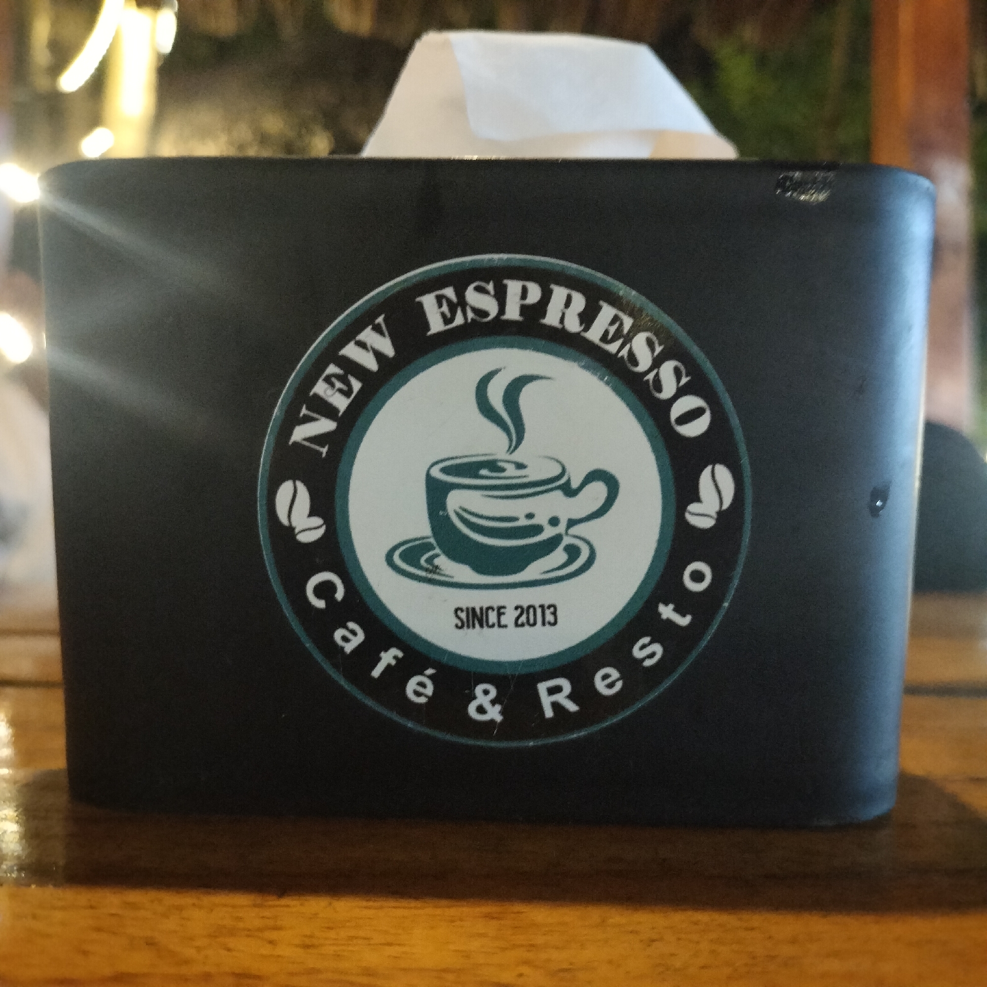 A close-up shot features a black tissue box with the logo of "New Espresso Café & Resto" prominently displayed. The logo is a circular design with the words "NEW ESPRESSO" along the top curve and "Café & Resto" along the bottom, separated by coffee bean icons. In the center is a stylized illustration of a coffee cup with steam rising from it, and the words "SINCE 2013" are below the cup.
A tissue is visible protruding from the top of the box. The box is placed on a glossy, wooden surface. Soft, blurred lights are visible in the background, suggesting an indoor or evening setting. The image was taken in Titibaro, Indonesia.