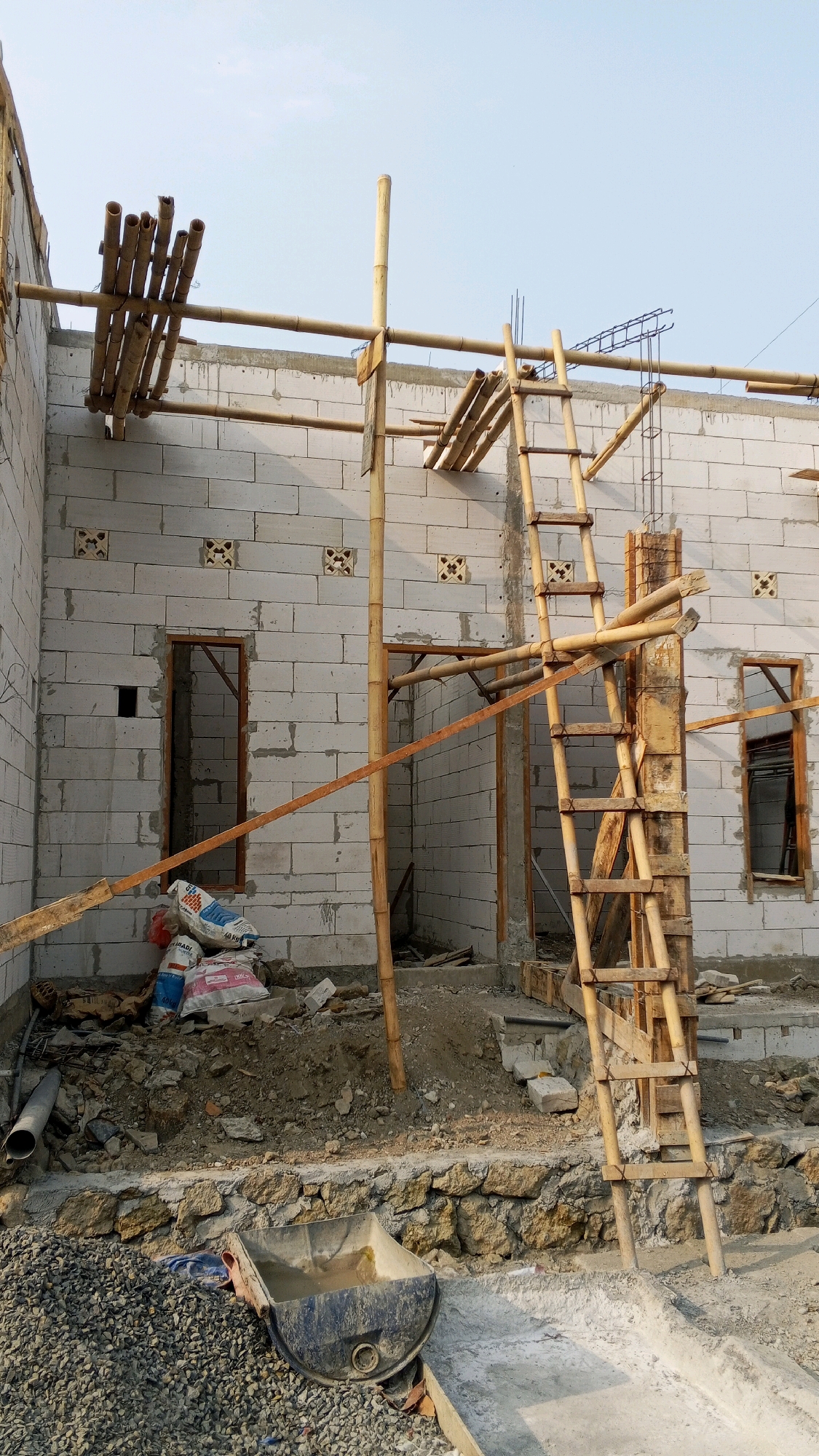 This image depicts an outdoor construction site for a building with white cinder block walls, likely in the city of Semarang, Indonesia. Scaffolding made of bamboo and wood supports a section of the roof structure, which appears to be under construction. Several stacks of cement bags are visible near the foundation, along with piles of dirt and gravel. A wooden ladder is leaning against the scaffolding. In the foreground, a dark blue plastic wheelbarrow filled with muddy water sits on a stone-lined embankment next to a pile of gravel. The sky is clear and bright, suggesting it is daytime. The overall scene is one of ongoing construction work.