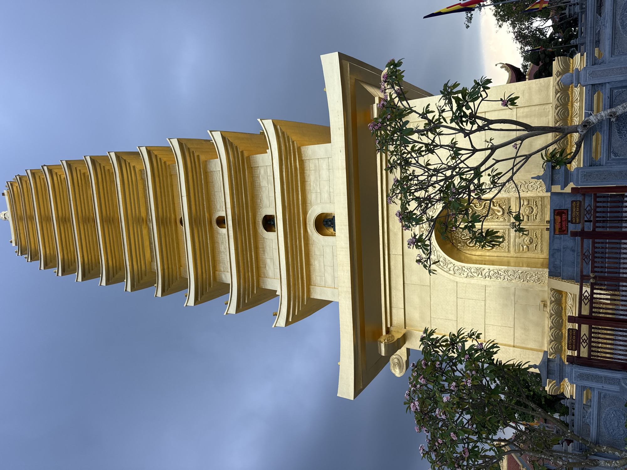 The image shows a tall, golden Buddhist pagoda in Diên Khánh, Vietnam. The pagoda has multiple tiers with curved roofs, and intricate golden detailing. In the foreground, there are two trees with pink flowers, and a stone fence. There are also flags visible to the right of the pagoda. The sky is cloudy and appears to be twilight or early evening. There are no people visible in the image.