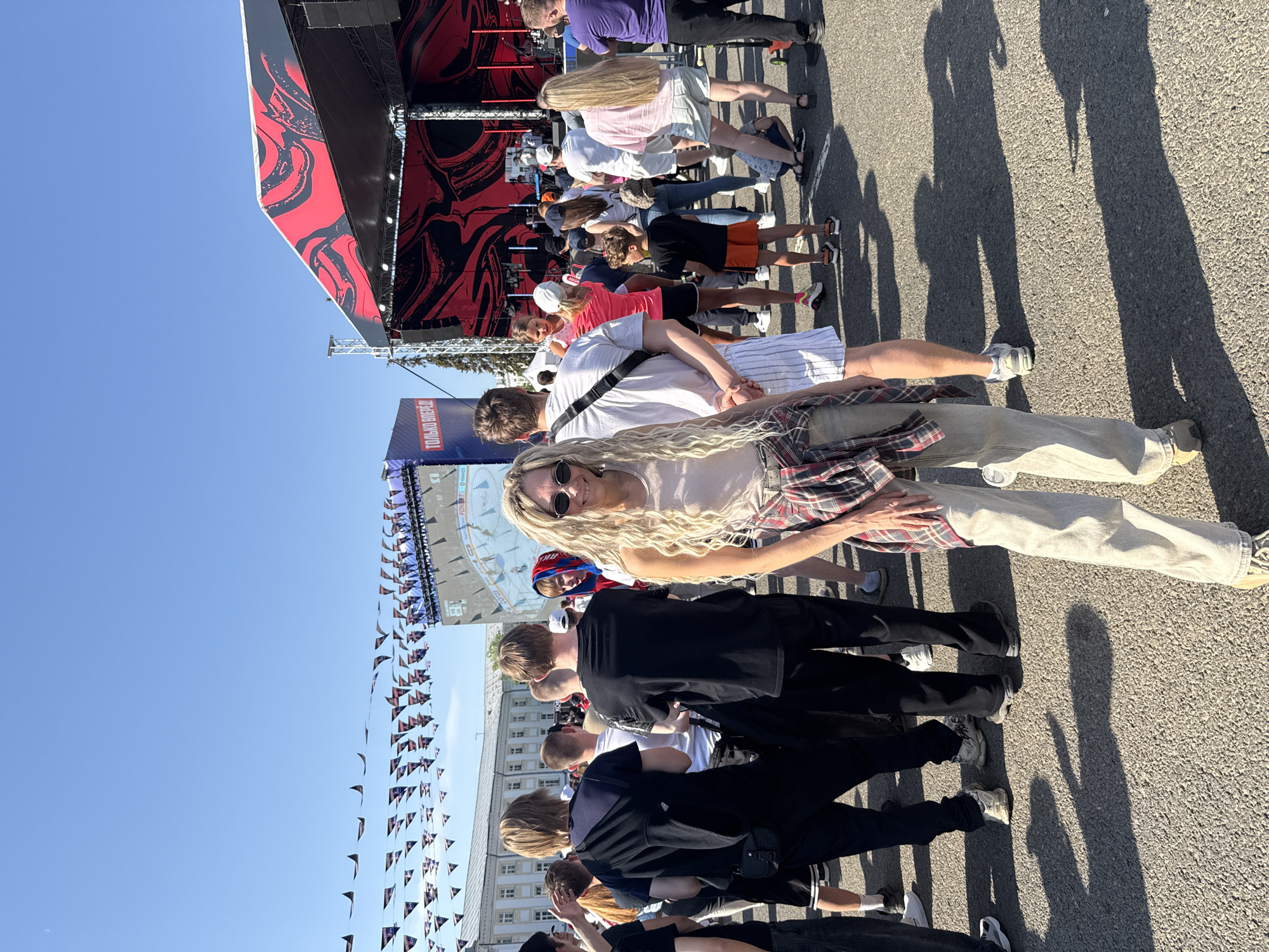 The image shows a crowd of people in Заволжье, Russia, possibly waiting for an event. A stage with red and black designs is visible in the background, along with banners of an ice rink. People are standing on an asphalt surface under a clear blue sky. Some individuals are looking at the camera. The atmosphere seems casual, indicating a public gathering or event.