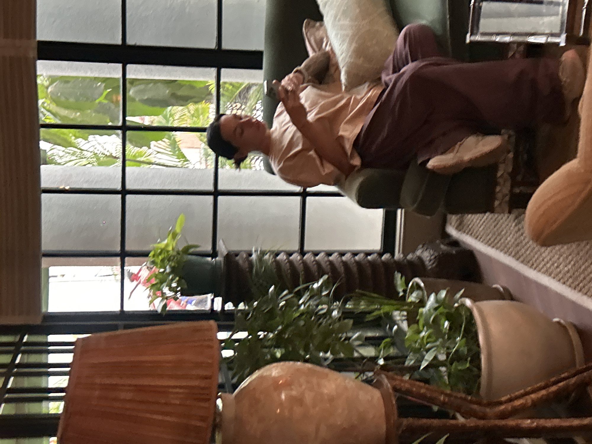 A person with dark hair pulled back is sitting on a dark green sofa in a well-lit room, holding a mobile phone and looking down at it. They are wearing a light pink t-shirt and loose-fitting dark purple pants. A light-colored throw pillow rests behind them on the sofa. The room features a large window with a grid pattern, revealing lush green foliage outside. In the foreground, to the left, is a large lamp with an orange pleated shade and a ceramic base. Various potted plants are visible, including one on a decorative stand. The flooring appears to be a patterned rug. The setting is indoors, and the overall ambiance suggests a relaxed atmosphere. This scene is located in Mumbai, India.