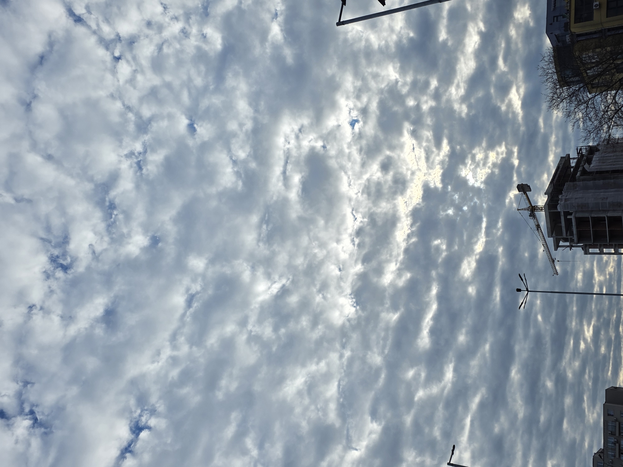 The image shows a view looking up at a cloudy sky in Plovdiv, Bulgaria. Visible in the lower right corner is a construction site with a crane. Streetlights are present throughout the scene. Buildings can be seen on the top and bottom edge of the image. The clouds are white and grey, with patches of blue sky visible in some areas. The sun appears to be shining through the clouds.