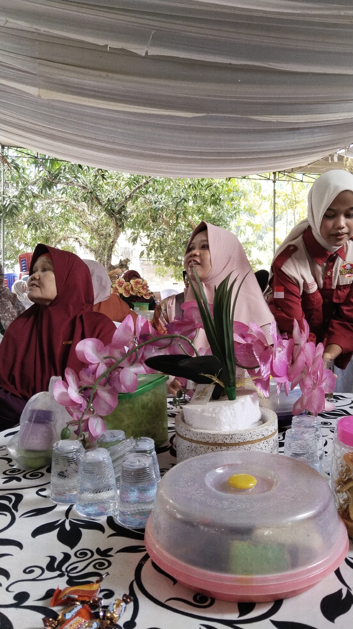 An outdoor gathering is taking place under a white tent. Three women are seated at a table covered with a black and white patterned cloth. The woman on the left wears a dark red hijab and a matching outfit. To her right, a woman in a light pink hijab is looking to her left, with a partial view of another woman in a white hijab and a red and white uniform on the far right. A floral arrangement with pink orchids is prominent on the table, along with several clear glasses filled with water, a green container, and wrapped candies. In the foreground, a pink and clear plastic container with a yellow lid sits on the table. The background reveals lush green trees and foliage, suggesting a natural, possibly residential, setting in Marga Sakti, Indonesia.