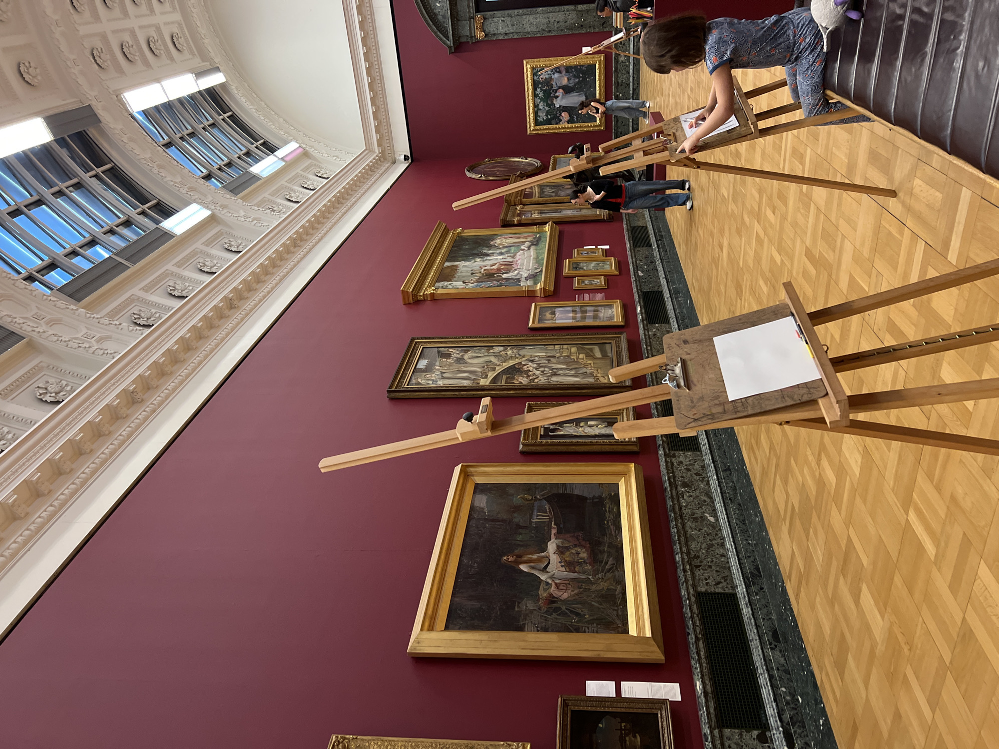 This media file captures an art activity taking place within a grand art gallery or museum, likely located in London, United Kingdom.
**Subjects:** The scene features several individuals engaged in drawing. A young child, dressed in a patterned blue outfit, is actively sketching on paper at a wooden easel in the foreground. Another person in a dark top and jeans is also drawing at an easel further down the gallery floor. Partially visible are other participants and attendees. The gallery walls are adorned with numerous framed paintings of various sizes, predominantly featuring gold or dark wooden frames. The floor is made of light-colored parquet wood. The ceiling is ornately decorated with white plaster moldings and features large arched windows that allow ample natural light into the space. A dark marble or stone baseboard with a metal grate runs along the bottom of the red wall.
**Activity or event taking place:** The primary activity is an art workshop or sketching session, where participants, including children, are set up with easels to draw. They appear to be observing and interpreting the artworks displayed on the walls.
**Notable details:** The setting is a formal art gallery, characterized by its deep red walls, classic framed paintings, and impressive, high ceilings with elaborate architectural details. The large windows suggest it is daytime. The atmosphere appears calm and focused, indicative of an educational or recreational art program. Small, illegible white labels are visible beneath some of the paintings, providing information about the artworks.