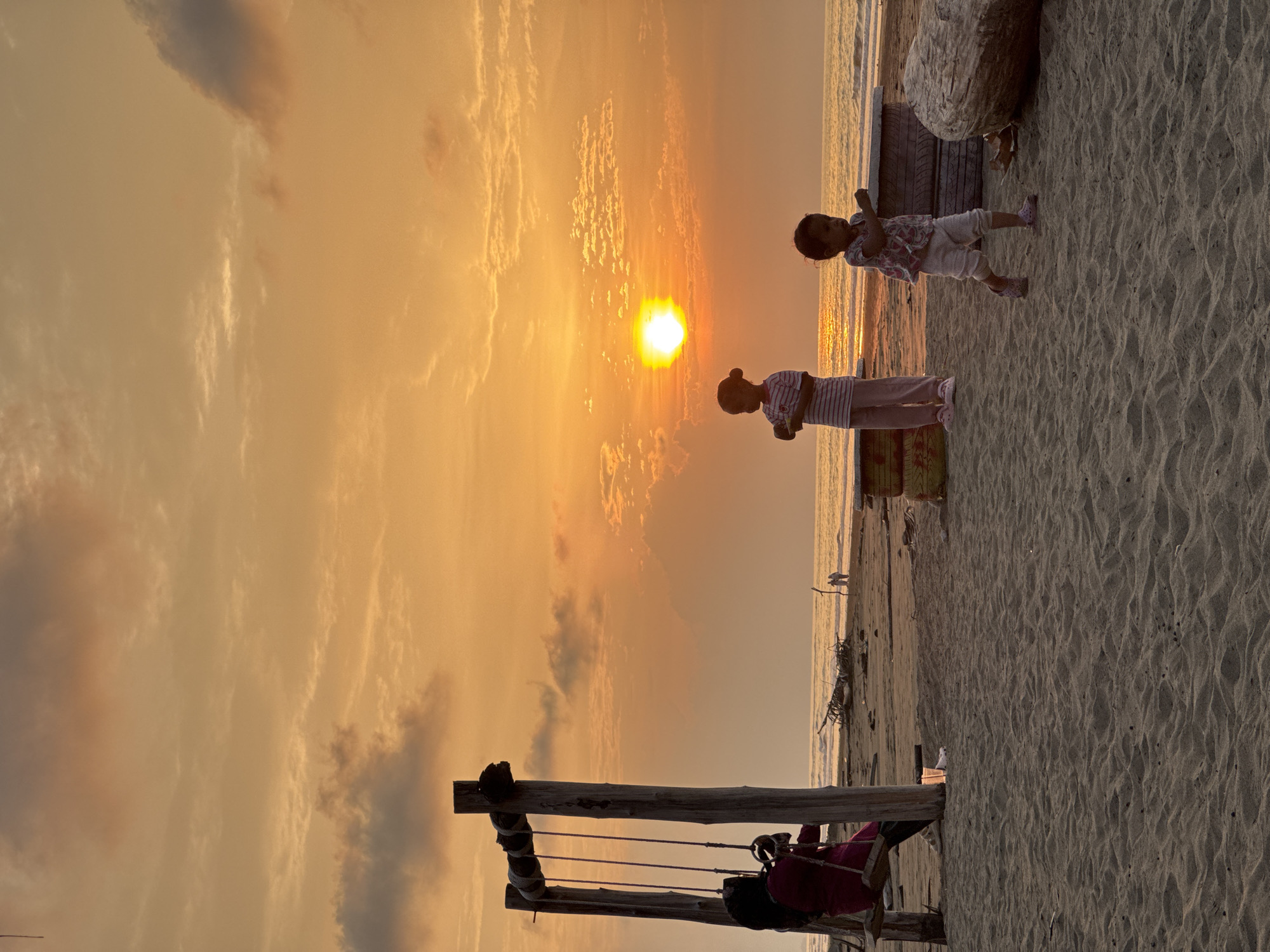 This image depicts a serene beach scene in Reudeuep, Indonesia, during sunset. The sky is awash in warm orange and yellow hues, with scattered clouds reflecting the fading sunlight. In the foreground, a sandy beach stretches towards the ocean.
On the left, a rustic wooden swing set stands with a person seated on it, wearing a dark headscarf and a maroon outfit. The person appears to be enjoying the view.
Further into the scene, two young children are present on the sand. One child, wearing a striped shirt and pink pants, stands facing left. The other child, dressed in a patterned top and light-colored pants, is captured in motion, walking towards the right.
Several objects are scattered on the beach, including what appear to be repurposed tires used as seating or decorative elements, and a large piece of driftwood. In the distance, the ocean waves gently lap the shore, and two silhouetted figures are visible walking near the water's edge. The overall atmosphere is peaceful and idyllic, capturing a moment of leisure and natural beauty.