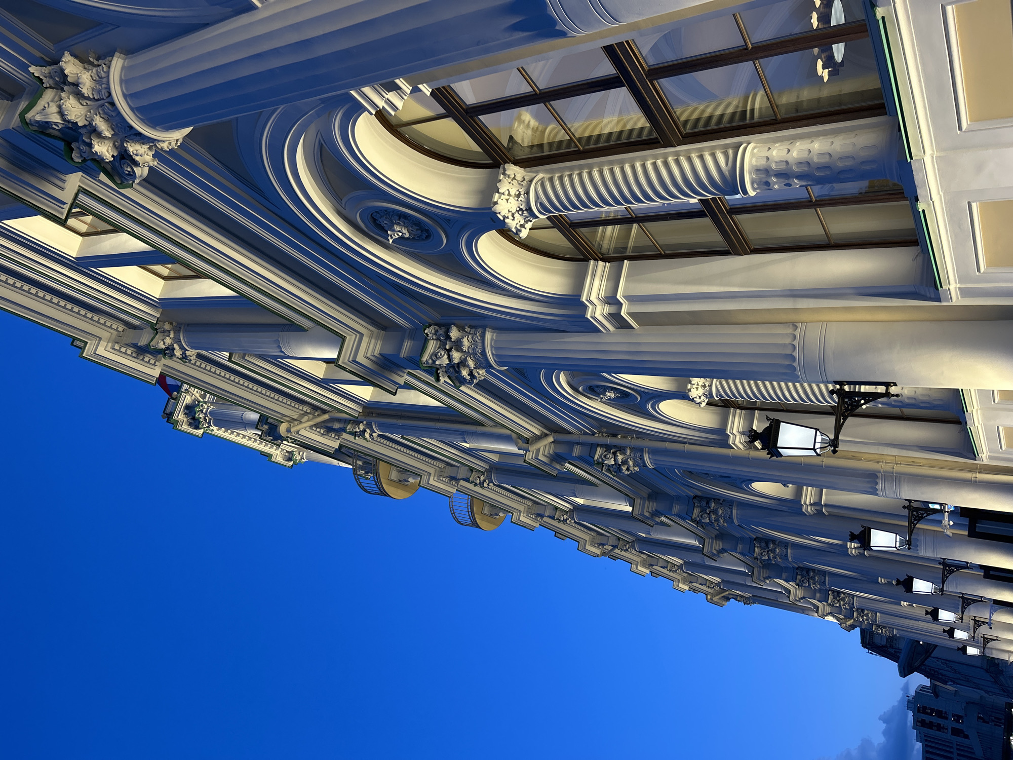An ornate architectural facade dominates the upper portion of the image, set against a deep blue sky that occupies the lower half. The building features a grand colonnade or a series of bays, adorned with numerous classical columns. These columns display intricate detailing; some have fluted shafts, while others exhibit spiral or ribbed patterns. Their capitals are elaborately carved in a style resembling Corinthian or Composite orders, rich with acanthus leaves and decorative volutes.
Between and above the columns, the building is embellished with a complex array of cornices, moldings, and arched windows. Some windows are large and rounded, featuring dark frames and reflecting light. The entire facade is brightly illuminated, creating a striking contrast with the darkening sky and highlighting the white and cream tones of the structure. This artificial lighting casts strong shadows, emphasizing the three-dimensional depth and decorative complexity of the architectural elements.
Several ornate black metal lanterns with glass panels are affixed to the facade, extending outwards. Some of these lanterns appear to be lit or reflecting light, adding to the evening ambiance. The sky, a vibrant, clear blue at the top, transitions to a darker, almost purple hue towards the bottom right, indicating twilight or early night. In the far bottom right corner, faint outlines of modern city buildings can be seen against the darker sky.
The image is captured from a low-angle perspective, looking up at the majestic building. There are no people visible, and no specific activity is taking place. No visible text is present.