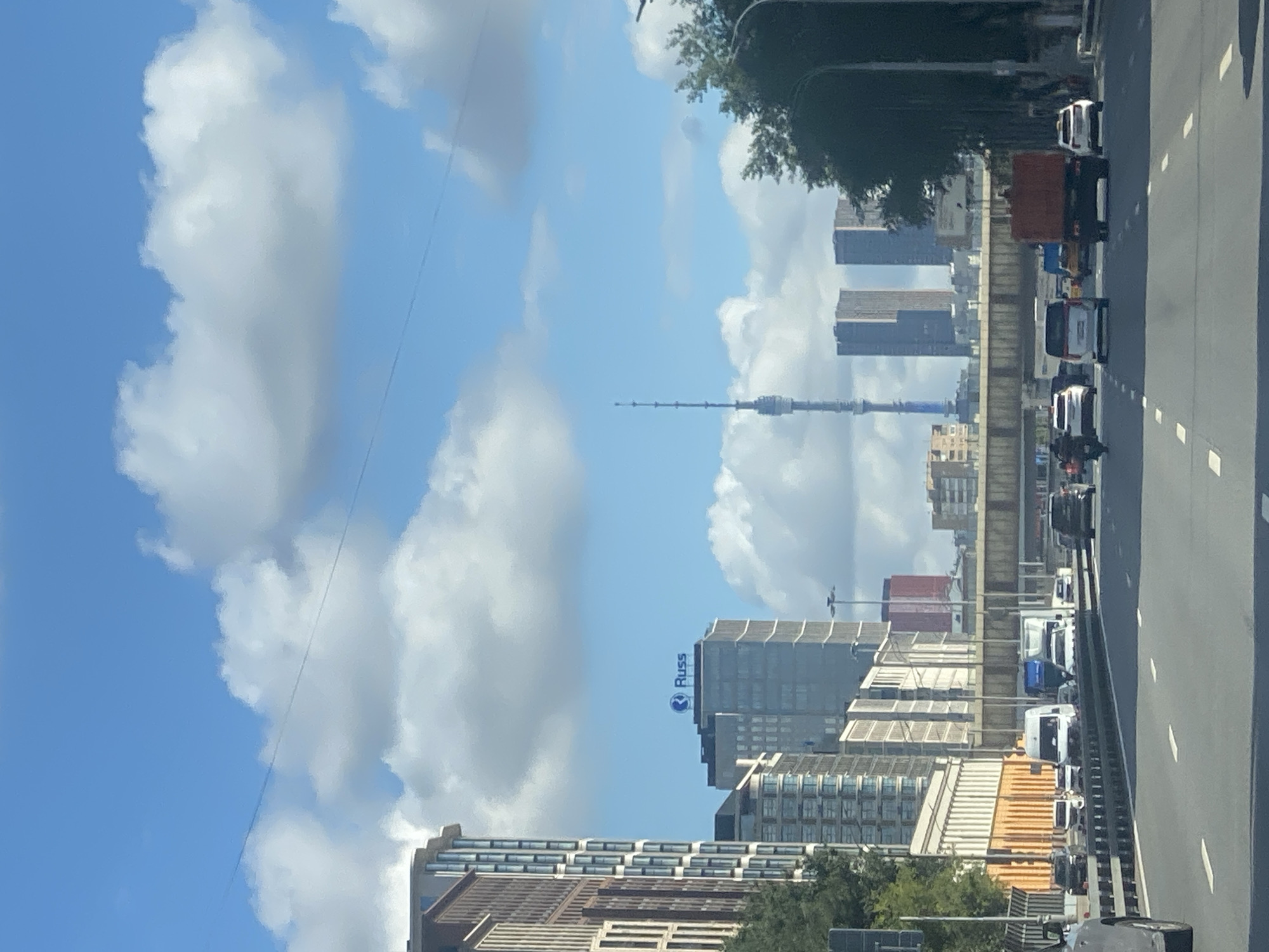 The image depicts a view from a highway in Moscow, Russia, on a day with a bright blue sky dotted with white clouds. In the foreground, several vehicles, including cars, vans, and a truck, are driving on the multi-lane road. The highway is bordered by a guardrail and trees. In the middle ground, a bridge spans over the highway, with more buildings visible behind it. Dominating the background is the Ostankino Tower, a prominent landmark in Moscow. To the left of the tower, several high-rise buildings can be seen, including one with a blue sign that reads "Russ". The overall scene suggests a busy urban environment with a mix of modern architecture and infrastructure.