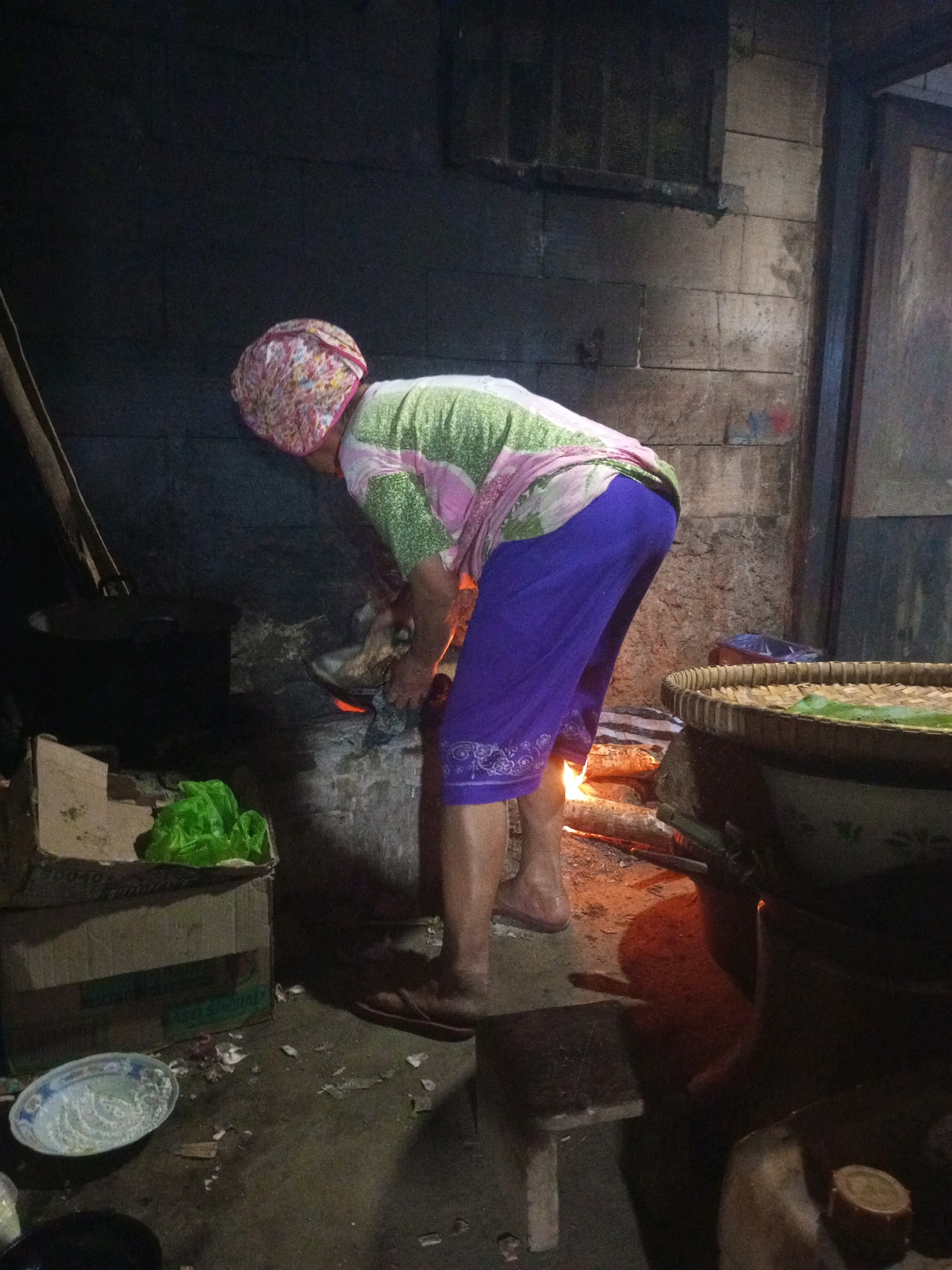 The image depicts an indoor scene, likely a kitchen, in Karangsari, Indonesia. A person, wearing a headscarf and a brightly colored patterned shirt with green and pink accents, is bent over a fire. They are wearing purple pants with a white embroidered pattern. The fire appears to be a traditional wood-burning stove. In the foreground, to the left, is a cardboard box with a green plastic bag inside and a blue and white patterned bowl on the ground. To the right, a large woven basket sits on top of a dark object, possibly another container. The walls are made of cinder blocks and appear weathered. The lighting is dim, with the fire providing a warm glow. The floor is dirt and scattered with debris.