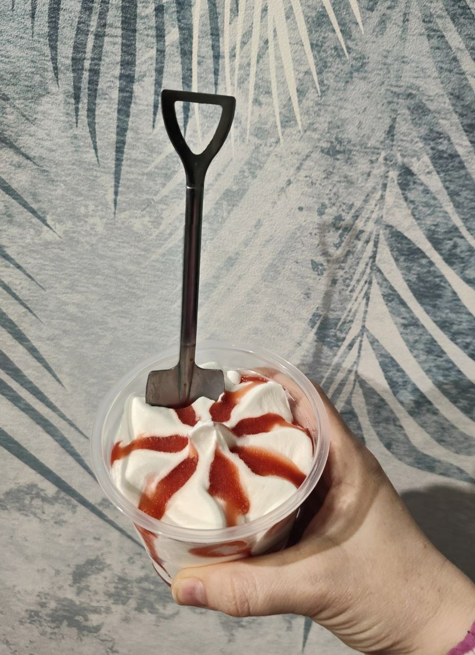 A hand holds a clear plastic cup of strawberry ice cream. A small, metal shovel is stuck into the center of the ice cream. The background is a wall with a pale blue and white leaf pattern. The image was taken in Minsk, Belarus.