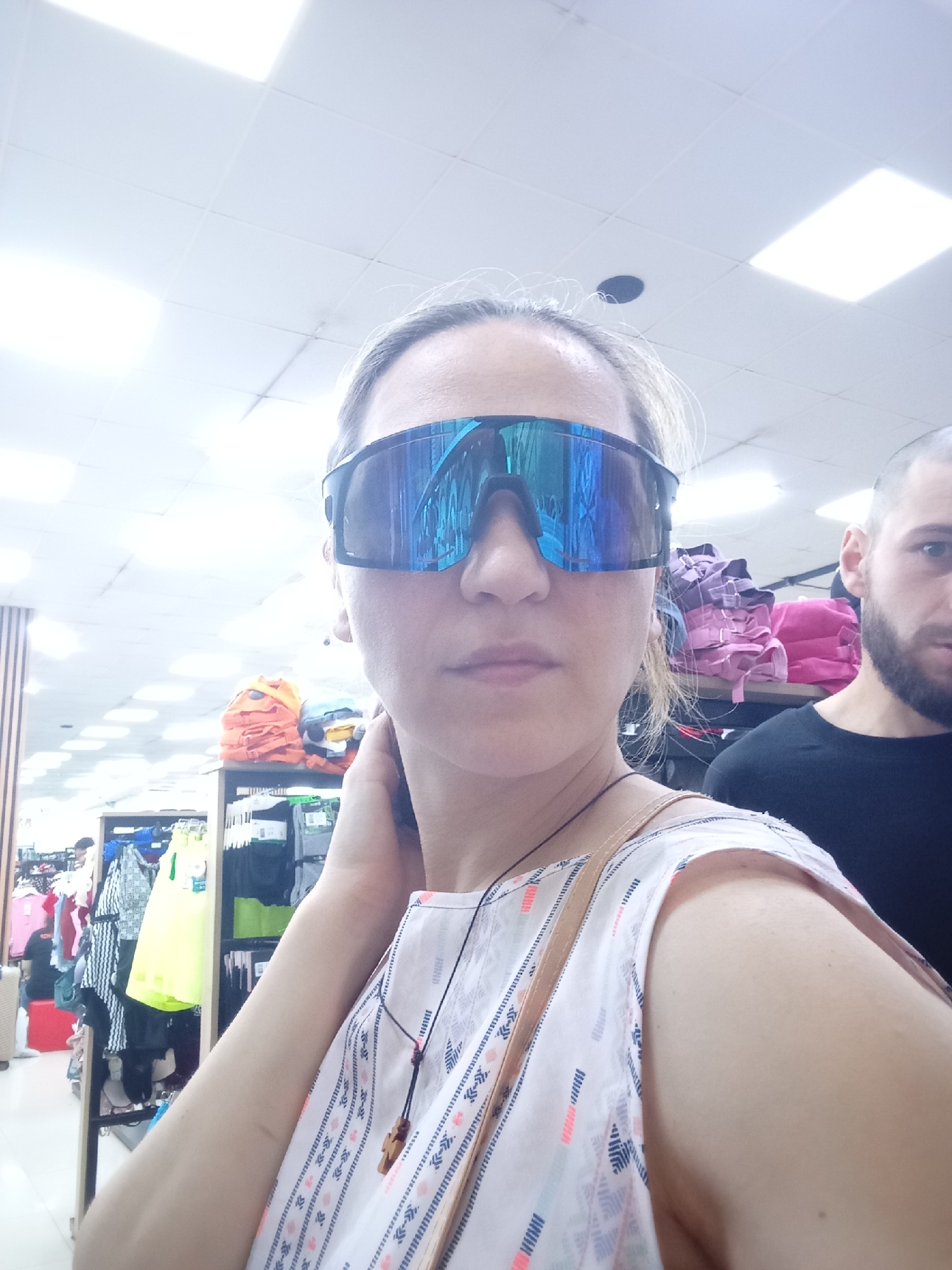 A woman wearing large, reflective, blue-tinted sunglasses and a patterned white top is taking a selfie in a store. She has her right hand near her ear and is looking towards the camera. A man with a beard and dark t-shirt is visible in the background to her right. The store appears to be a retail establishment with clothing racks and shelves stocked with merchandise, including folded fabrics and what appear to be bags. The lighting suggests an indoor commercial setting. The image was geotagged to Tbilisi, Georgia.