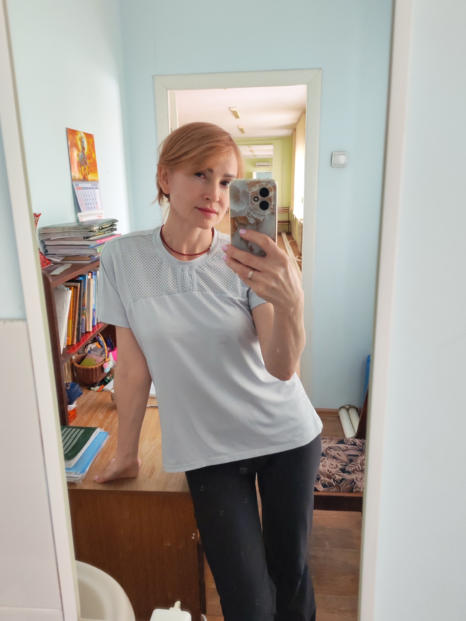 The image shows a woman taking a mirror selfie in a room located in the city of Chaikovsky, Russia. The woman, with short reddish-blonde hair, wears a light grey short-sleeved t-shirt with a mesh upper chest pattern, dark pants, and a thin red choker necklace. A gold-colored ring is present on her left ring finger, and her fingernails and visible toenails are painted light pink. She holds a smartphone with a floral-patterned case in her right hand, directed at the mirror, while her left forearm and bare left foot rest on a wooden desk or cabinet. The room has light blue walls. To the left, a wooden shelving unit contains stacked papers, books, and a small woven basket. A calendar featuring a white horse is hung on the wall above the shelves. A white light switch is visible on the wall to the right of the woman's reflection. Through an open doorway in the background, a light yellow hallway is visible, featuring fluorescent lighting, a patterned upholstered bench, and an air conditioning unit in the distance. Two white cylindrical objects lie on the floor near the doorway entrance.