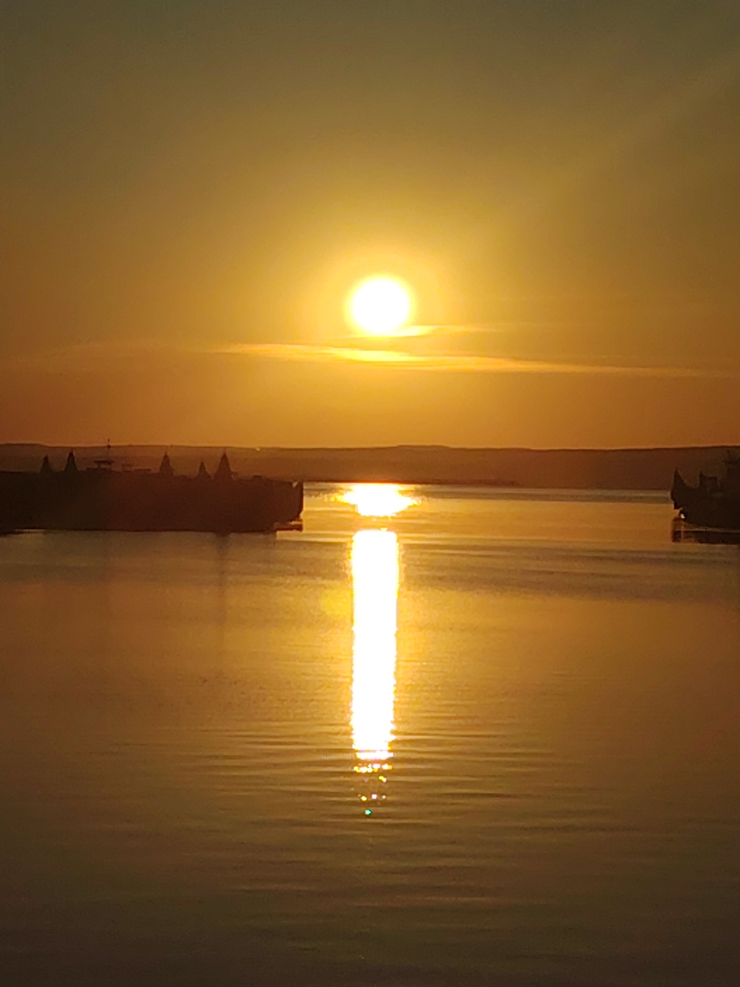 A wide, still body of water, likely the Kama River, is shown at sunset. The sun, a bright orb casting a golden glow, is positioned high in the orange sky, with subtle clouds visible. A dark, silhouetted structure, possibly a dam or a fortified structure with multiple spires, is situated on the left bank of the river, its reflection creating a shimmering path on the water's surface. The water is calm, with gentle ripples reflecting the sunset. The overall atmosphere is serene and tranquil. The location is identified as Chaikovsky, Russia.