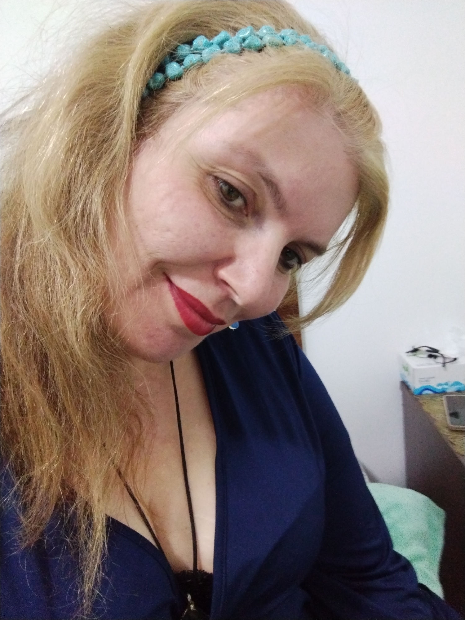 A woman is pictured indoors, wearing a blue top and a turquoise beaded headband. She is looking down and smiling with red lipstick. A thin black cord or necklace is visible. Behind her is a white wall, a dark wooden cabinet, a box of tissues, glasses, and a mobile phone. The location is not explicitly discernable as Al Mas'oudi, United Arab Emirates without additional identifying information within the image.
