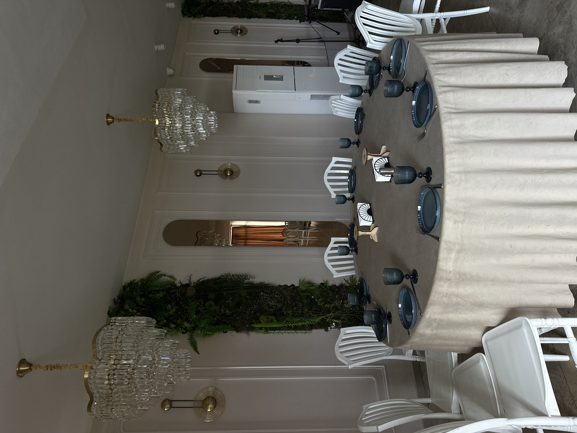 The image captures an indoor dining setup. A long, draped table occupies the central space, set with plates, glasses, and silverware. White chairs are arranged around the table. Above, ornate chandeliers hang from the ceiling. Decorations of greenery adorn the walls. A tall, white appliance stands against the wall in the background. The scene appears to be in Marneuli, Georgia, based on visible text on a sign.