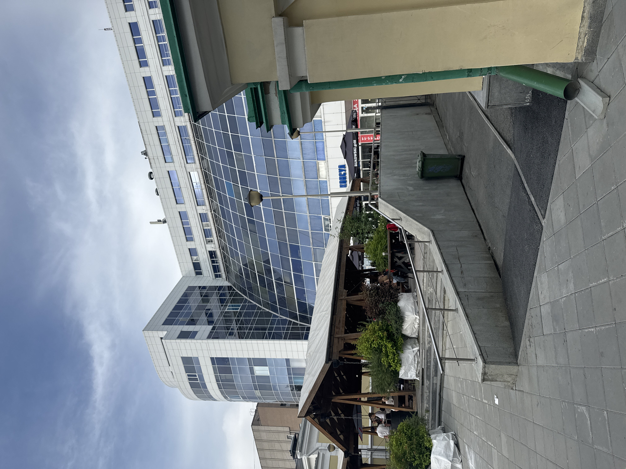 The image depicts an outdoor seating area adjacent to a modern glass-fronted building. The scene includes a covered wooden structure with tables and chairs, where several people appear to be seated. Lush green bushes are planted around the seating area, some covered in white fabric, possibly for protection or storage. A metal railing runs alongside a set of stairs leading to the area.
In the background, a large, contemporary building with a curved facade composed of glass panels and white framing dominates the view. Another, older building with a lighter facade is partially visible to the left. A green trash bin is situated on a paved pathway near the building.
The sky is overcast with visible clouds, suggesting a cloudy day. The orientation of the image is vertical, with the top of the buildings appearing on the left side of the frame.
Text visible on signage behind the covered seating area includes "BUKOWSKI Grill" and "127-11-11". The overall atmosphere seems relaxed and casual, likely a cafe or restaurant seating area during daylight hours. The location is identified as Yekaterinburg, Russia.