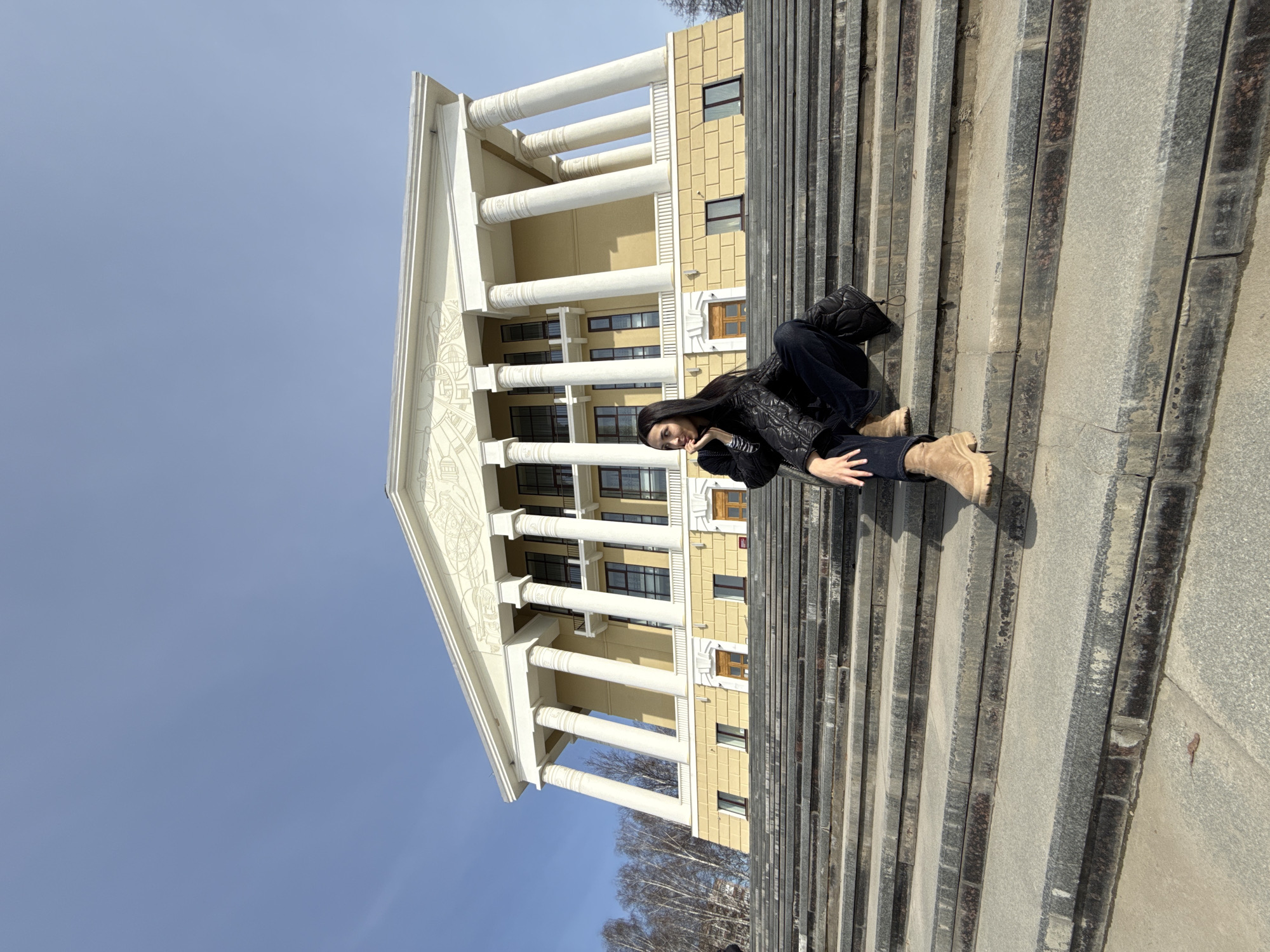 A woman is sitting on a set of granite stairs in front of the Tchaikovsky House Museum in Votkinsk, Russia. The building is a three-story beige structure with white columns and a triangular pediment. The sky is clear and blue. The woman is wearing a black jacket, dark pants, and tan boots. She is smiling and holding her hand to her face. A black bag is nearby on the steps. The stairs are granite and lead up to the museum entrance. Trees are visible in the background.