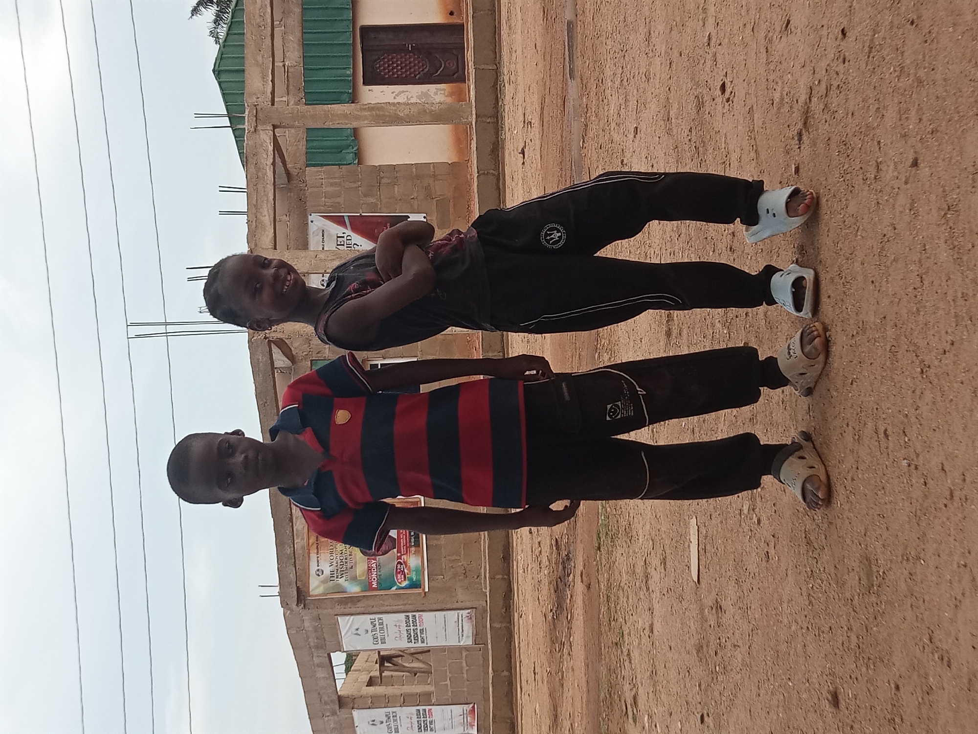Two children pose for a picture in Sabon Tasha, Nigeria. The child in the foreground wears a red and navy striped polo shirt, black pants, and tan sandals. Behind them, a child wears a black shirt with a red design, black pants, and white sandals, posing with their arms crossed. They are standing on a dirt ground next to a building under construction with visible posters. The background shows power lines against a cloudy sky.