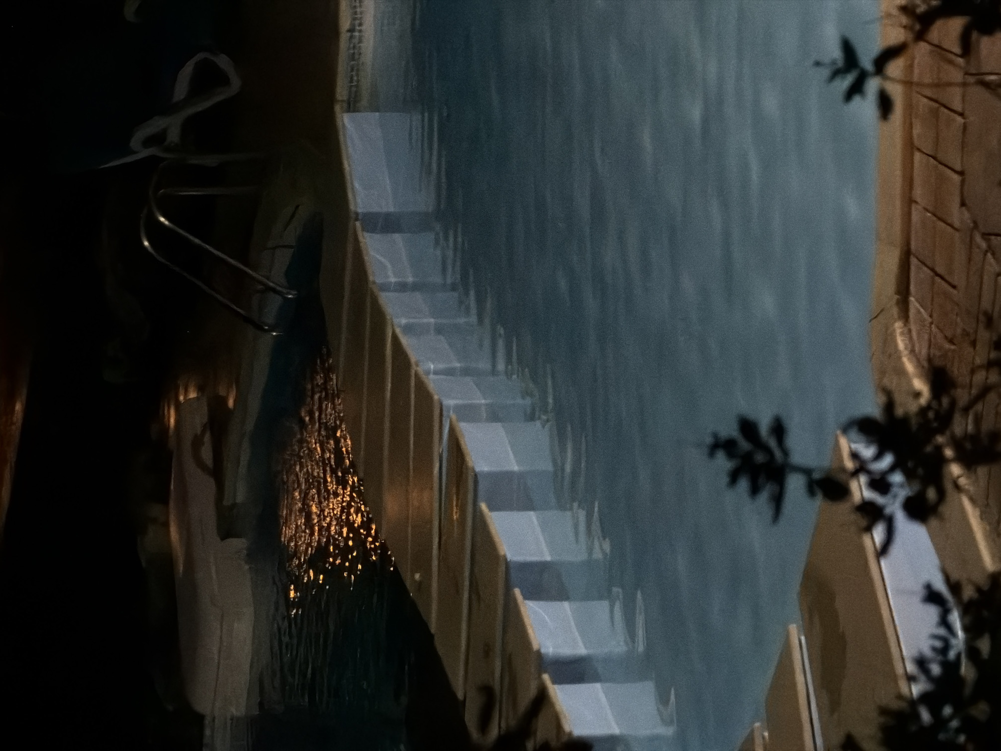 At night, a swimming pool is lit by artificial lighting, casting warm reflections on the water's surface. A wooden walkway with white supports extends across the pool, creating a pathway over the water. In the background, a metal ladder with a curved handle suggests an entrance to the pool. The surrounding area is dark, with some blurry shapes indicating foliage or structures. The scene appears to be an outdoor recreational area, possibly a resort or private residence. This image does not contain any information to specify the location as Sharm El Sheikh, Egypt.