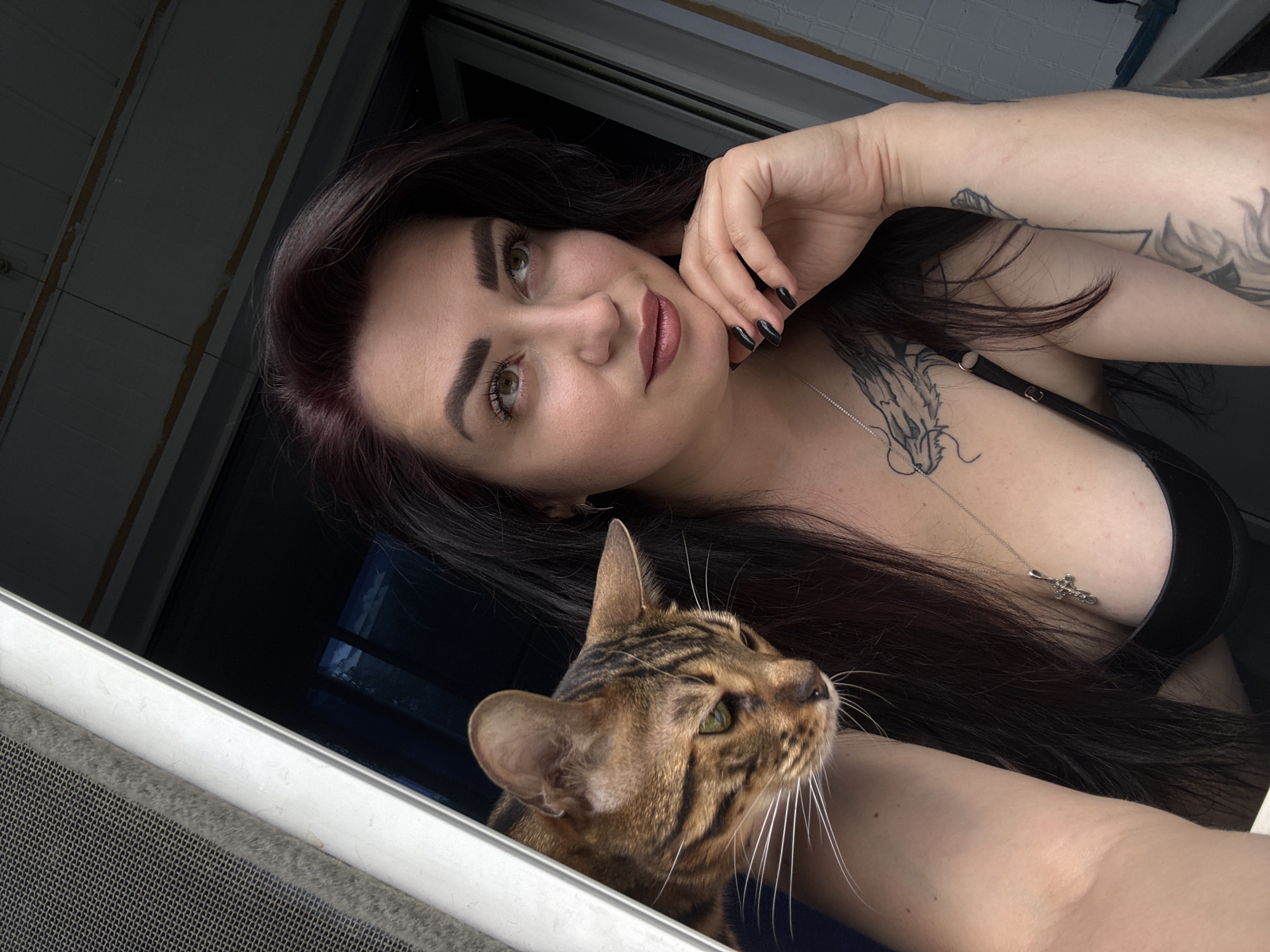 In a medium shot, a woman with dark hair and tan skin poses indoors. She is looking to her right, with her chin resting on her left hand, which is positioned near her mouth. The woman is wearing a black top and a silver necklace with a cross pendant. She has tattoos on her chest and left arm. Next to her, a tabby cat peeks into the frame from the left, its head and shoulders visible. The cat's body is partially obscured by the woman's arm. The background appears to be a window or doorway, with a screen visible on the left side. The lighting suggests an indoor setting with natural light coming from the outside. The location is listed as Unknown, Unknown.
