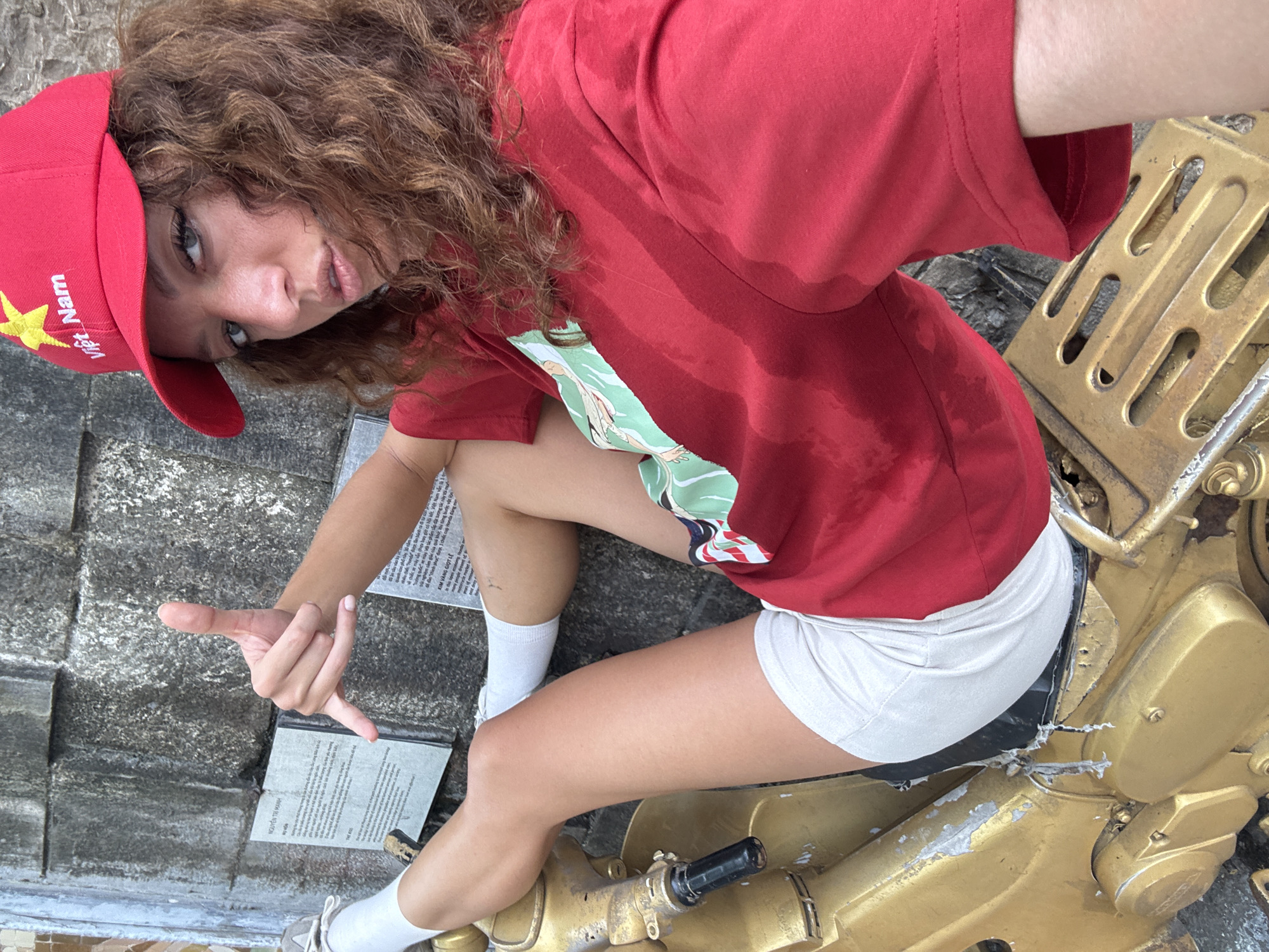 A young woman with curly brown hair, wearing a red t-shirt, white shorts, and a red "Việt Nam" cap, is sitting on a large, gold-colored artillery weapon. She is making a hand gesture resembling the letter "L." The photo is taken from a high angle. Two stone plaques containing text are visible near the woman. The location is in Hà Nội, Vietnam.