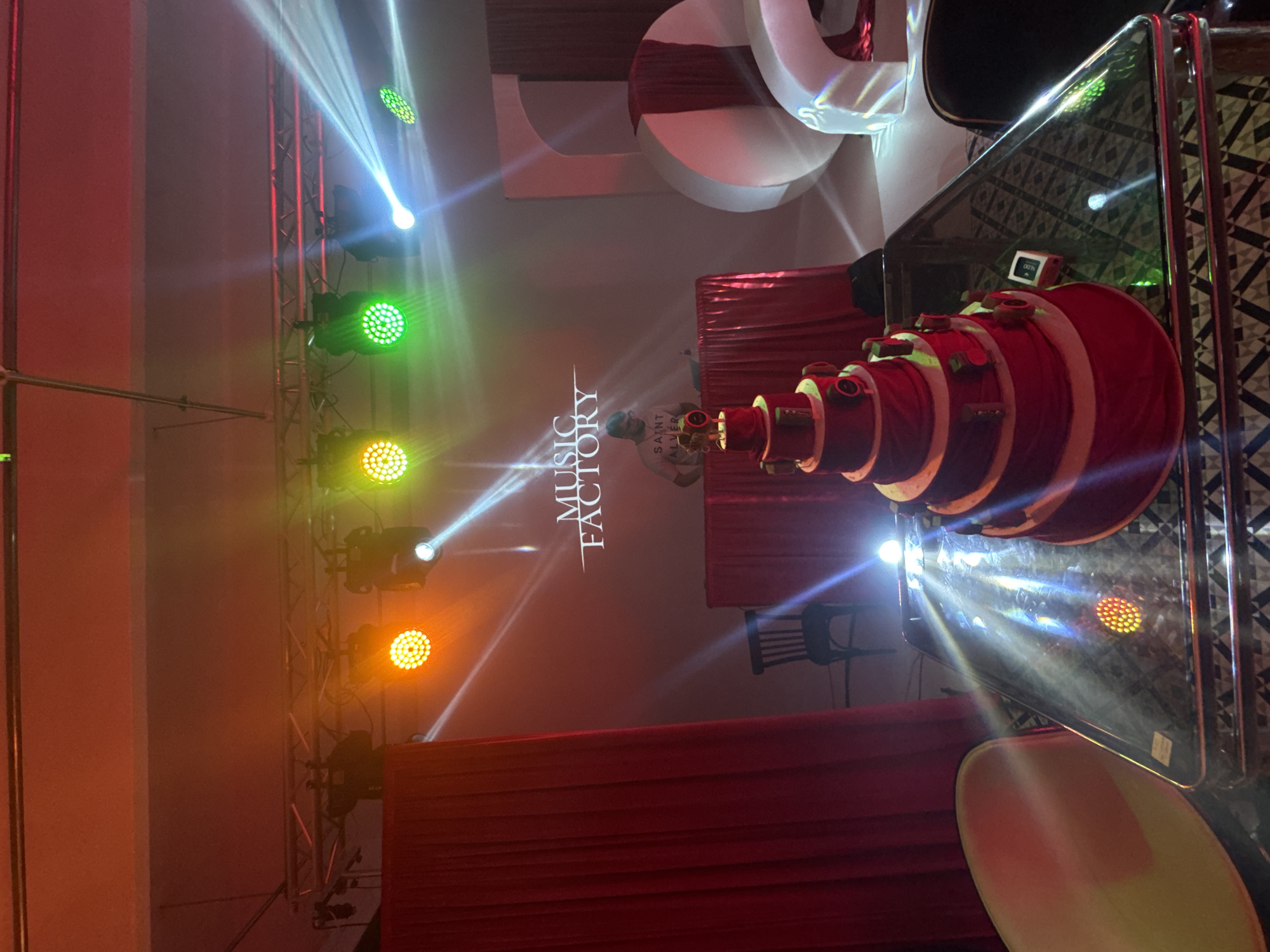 The image shows an indoor event venue, likely a club or stage, possibly in Busmantsi, Bulgaria. Red curtains are draped in the foreground and background. In the center, there is a projection of the words "MUSIC FACTORY" along with a human figure. Stage lighting rigs are visible on the left side of the frame, with various lights emitting beams of different colors (white, green, orange). To the right of the stage projection, there is a circular, layered structure, predominantly red and white, positioned near what appears to be a table with a patterned surface. A chair is partly visible near the table. The overall ambiance is one of a performance or event.