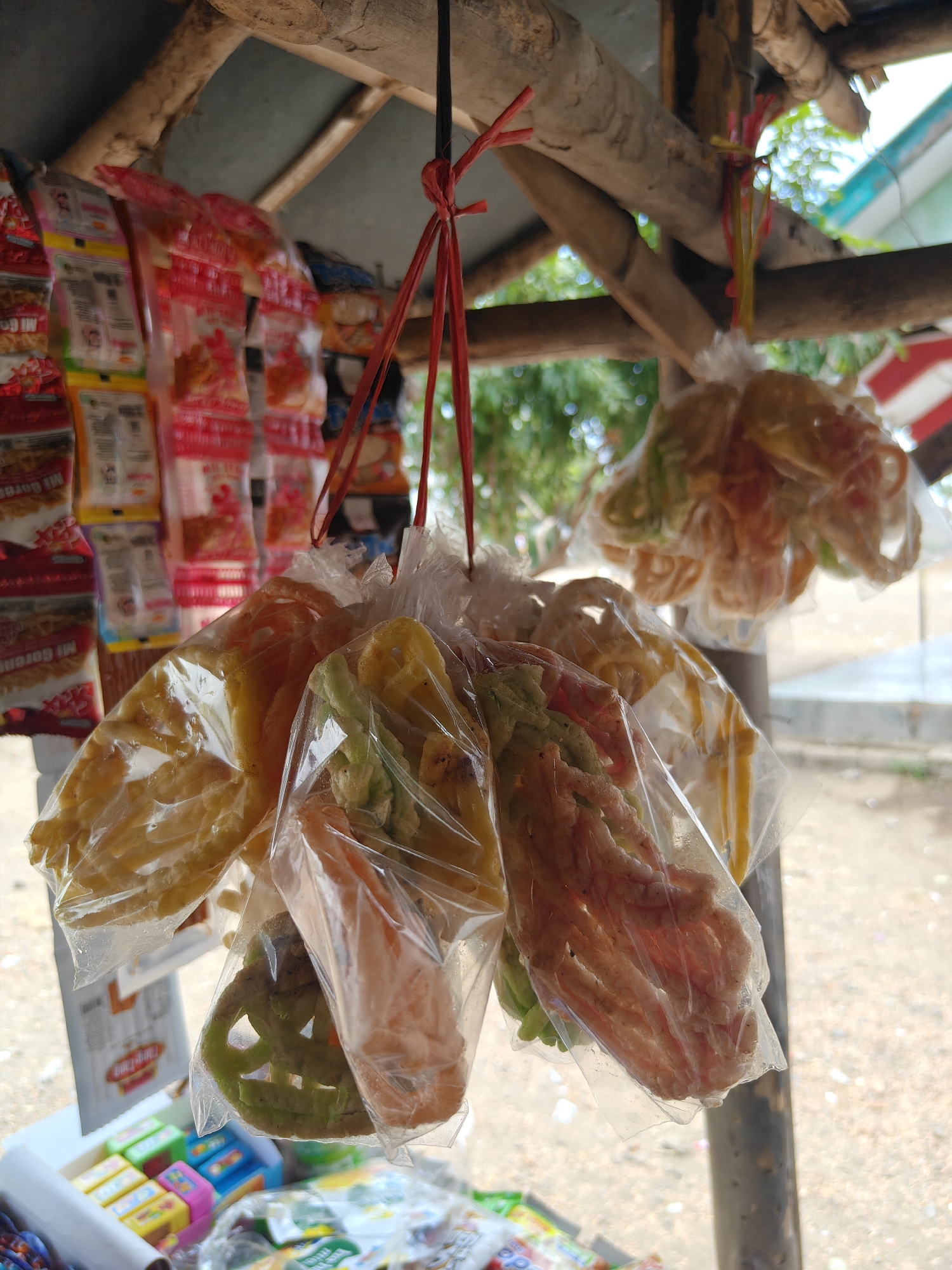 The image depicts an outdoor stall in Cermee, Indonesia, displaying an assortment of packaged snacks and colorful fried snacks hanging from a rustic wooden structure. In the foreground, several plastic-wrapped bundles of these fried snacks are prominently displayed. These snacks vary in color, appearing in shades of yellow, orange, and green, and have intricate, wavy shapes. Behind these, on the left side of the frame, rows of commercially packaged snacks, predominantly featuring red and yellow branding, are neatly arranged on a shelf. The stall itself appears to be constructed from natural wood, with a rudimentary roof overhead. The background reveals a blurred outdoor environment with hints of greenery, suggesting a natural or semi-rural setting. A white surface, possibly a counter or display box, is visible at the bottom, with some smaller colorful items, possibly candy or more snacks, arranged on it. The overall scene conveys a sense of a local, informal market or roadside vendor.
