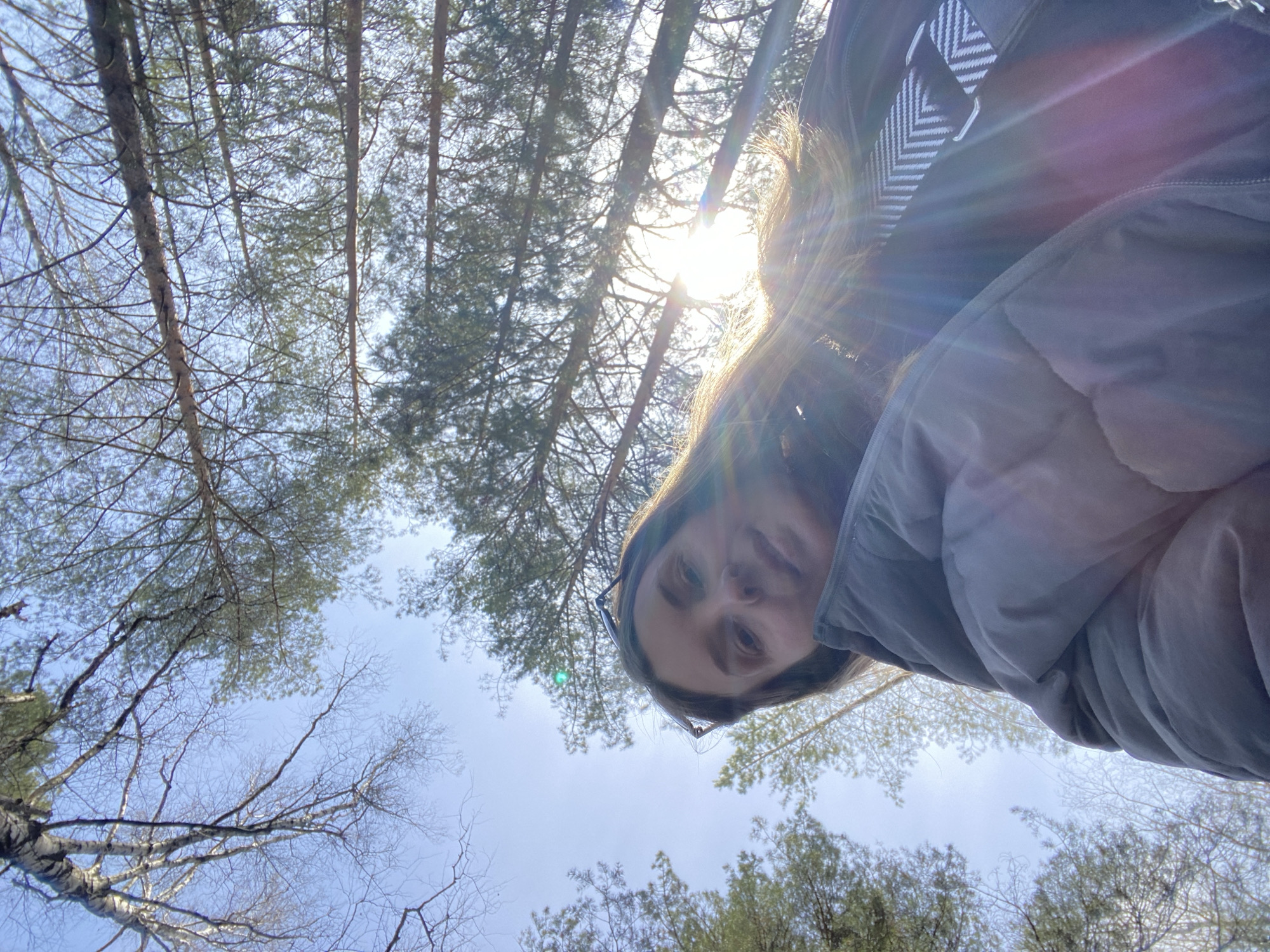 A low-angle shot looking upward shows a person in a puffy jacket standing in what appears to be a forest or wooded area in Noviy, Russia. The trees, including some with bare branches, fill much of the frame. The sky is partially visible through the trees. The sun is obscured by the trees, but its presence creates a bright glare. The person in the jacket is looking down into the camera. A portion of their hair and glasses are visible.
