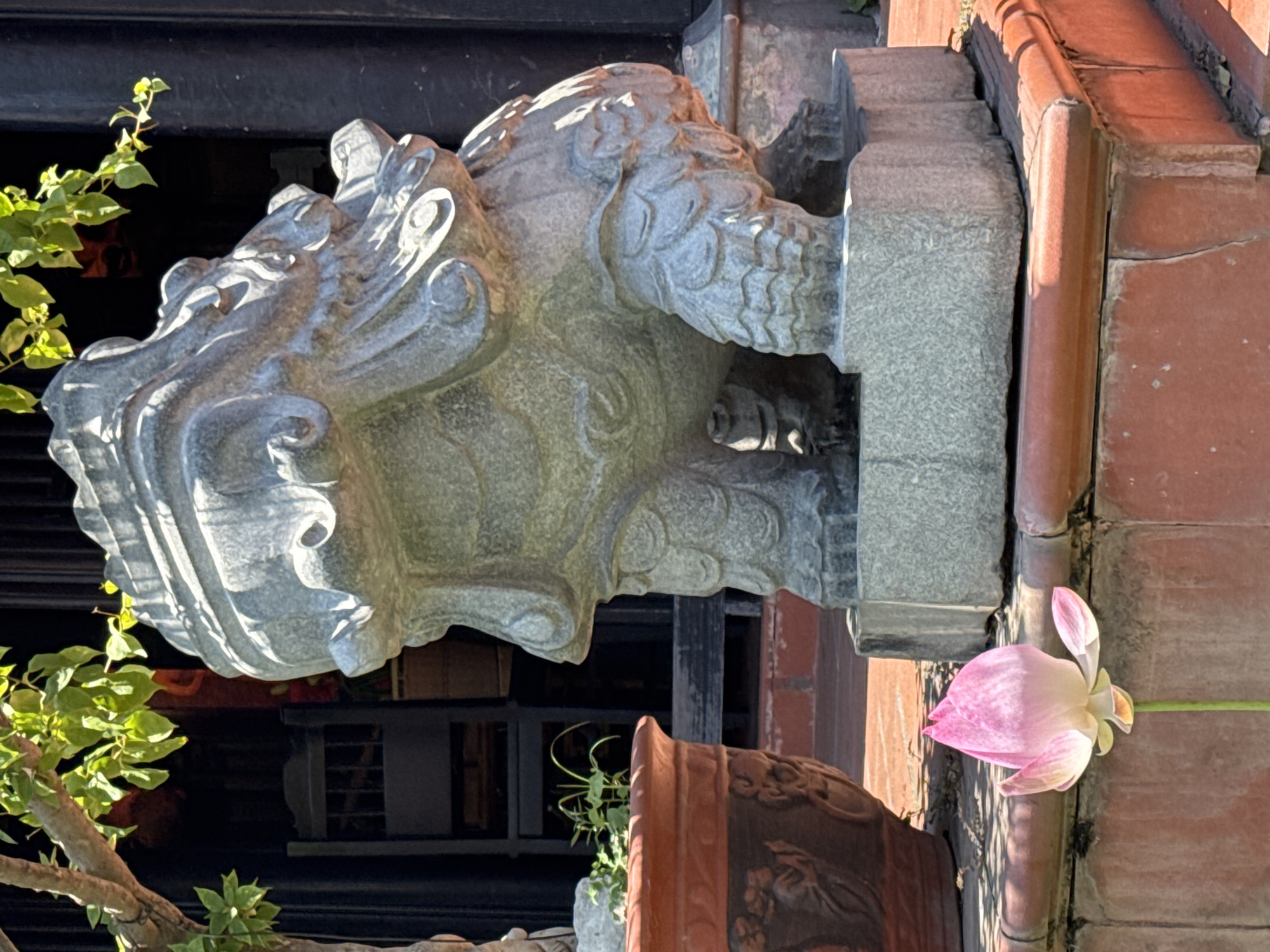 A stone lion statue is positioned adjacent to a red-tiled wall with a curved edge, likely a roof corner or decorative element. A pink lotus flower is situated near the wall, seemingly growing from or attached to it. A leafy branch extends from the left side of the frame. Behind the statue, the facade of a building is visible, appearing dark. The setting may be in Bac Nha Trang Ward, Vietnam.