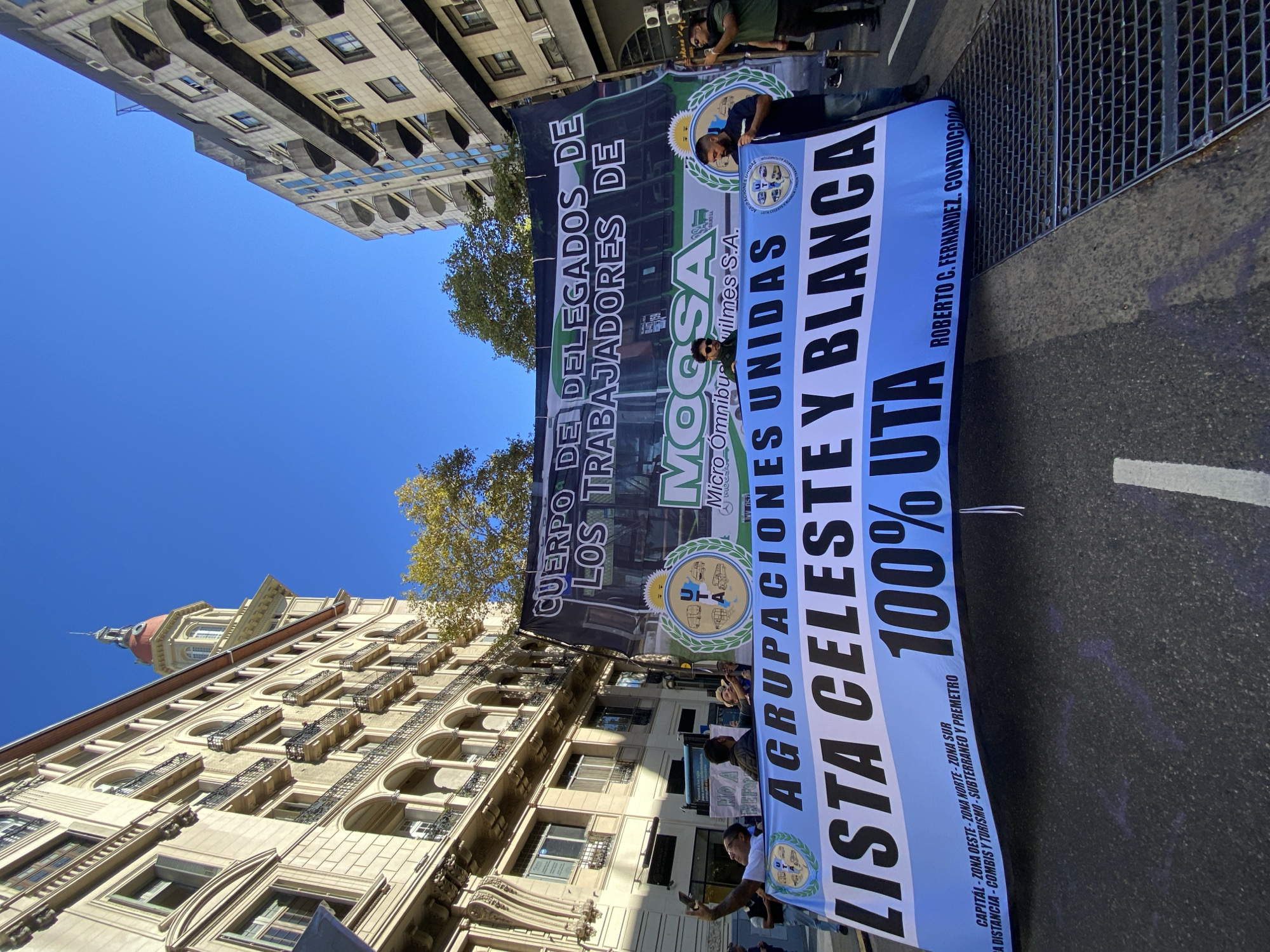This image captures a vibrant street scene in Buenos Aires, Argentina, on a clear, sunny day, likely during a protest or public demonstration. The picture is rotated, showing a low-angle perspective looking up towards tall buildings and a bright blue sky.
The main subjects are several individuals, mostly men, holding two large banners stretched across the street. The banners are the focal point of the event, indicating a workers' gathering.
The larger, darker banner at the back reads "CUERPO DE DELEGADOS DE LOS TRABAJADORES DE" (BODY OF DELEGATES OF THE WORKERS OF). Below this, the logo for "MOGSA Micro Ómnibus Quilmes S.A." (a bus company) is visible.
The front banner is white and light blue, prominently displaying "AGRUPACIONES UNIDAS" (UNITED GROUPS) and "LISTA CELESTE Y BLANCA" (LIGHT BLUE AND WHITE LIST), which are common political colors in Argentina. It also states "100% UTA," referring to the "Unión Tranviarios Automotor" (Automotive Tramway Union), a transport workers' union. The name "ROBERTO C. FERNANDEZ, CONDUCCIÓN" (Roberto C. Fernandez, Leadership) is also present, likely indicating the union's leader. Further text on this banner details the union's operational areas: "CAPITAL - ZONA OESTE - ZONA NORTE - ZONA SUR" (Capital - West Zone - North Zone - South Zone) and services such as "A DISTANCIA - COMBIS Y TURISMO - SUBTERRANEO Y PREMETRO" (Long Distance - Vans and Tourism - Subway and Premetro).
The setting is an urban street lined with multi-story buildings featuring classical architecture, balconies, and some air conditioning units. A glimpse of a tree with green foliage is visible. The bright sunlight suggests a daytime event. On the ground, parts of a street, including grates and painted lines, are visible.