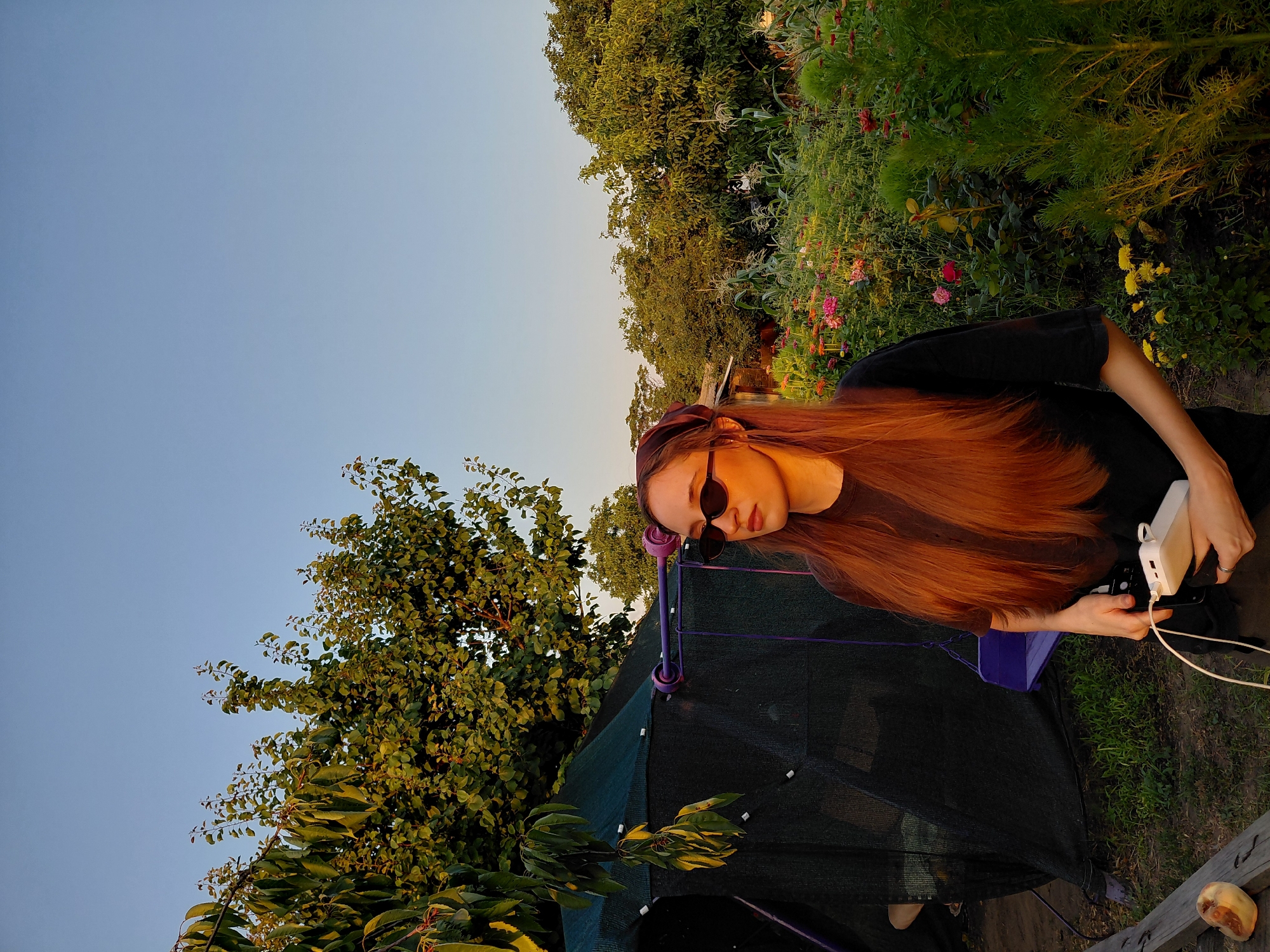 A young woman with long, auburn hair and sunglasses is standing in a garden at sunset. She is wearing a dark t-shirt and black pants. In her hands, she holds a white portable charger connected to a cable. Behind her is a large tree with green leaves, a black net structure with purple poles and a purple swing, and a vibrant garden filled with various flowers and green foliage. The sky is a clear, pale blue, indicative of late afternoon or early evening light.
