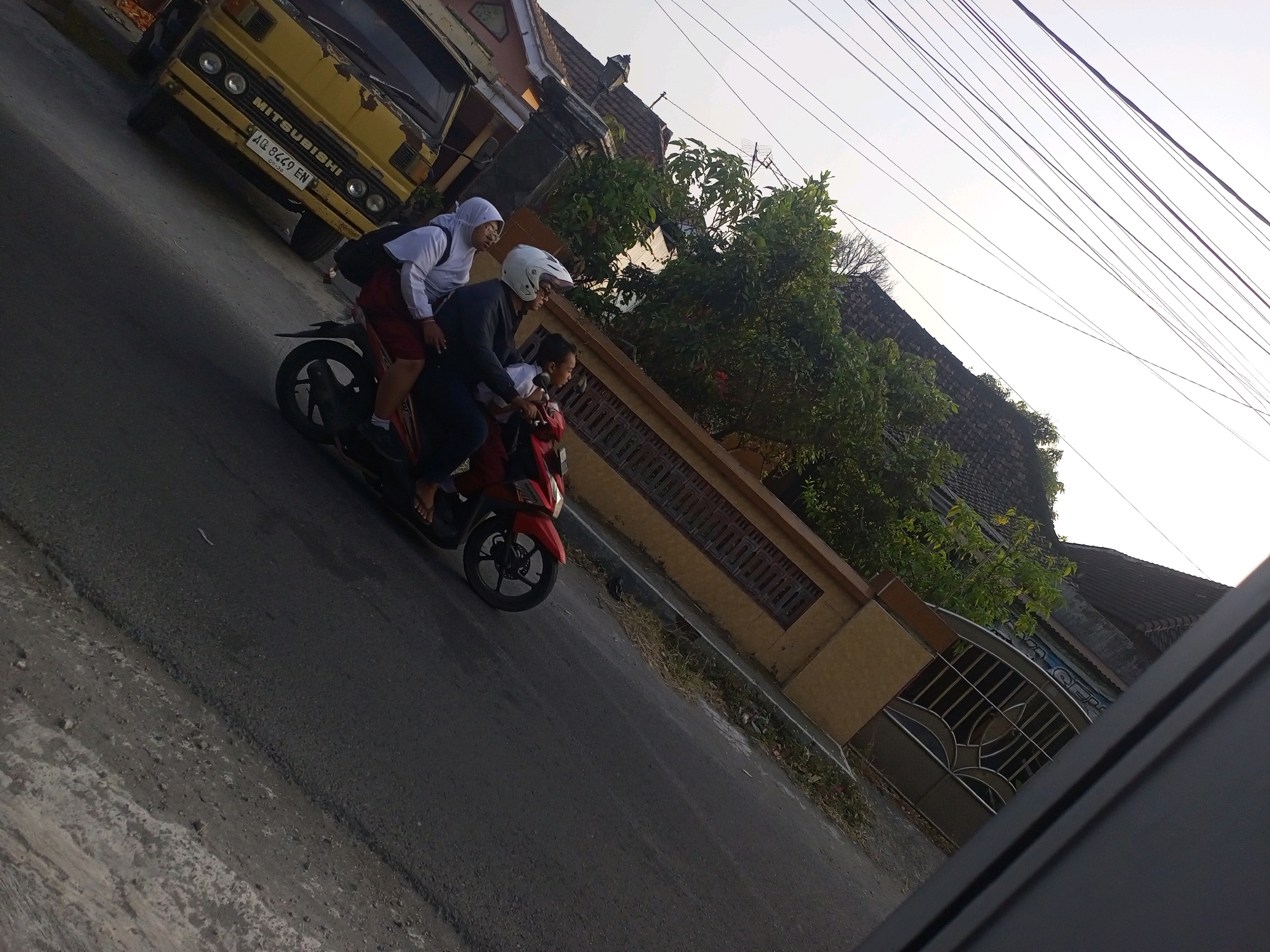 In the city of Ngasem, Indonesia, on a paved road, three individuals are riding on a red scooter. A person in a white hijab and red skirt is seated in the middle, holding onto the person in front. The person in the middle is wearing a white hijab and a red skirt. In front of them, a person wearing a white helmet and dark clothing is driving. A child is seated behind the driver, facing forward. The scooter is moving past a yellow Mitsubishi truck parked on the left side of the road, which displays a license plate with the characters "AQ 8469 EN". The background features houses with tiled roofs and lush green trees. The sky is overcast with power lines crisscrossing the upper portion of the frame.