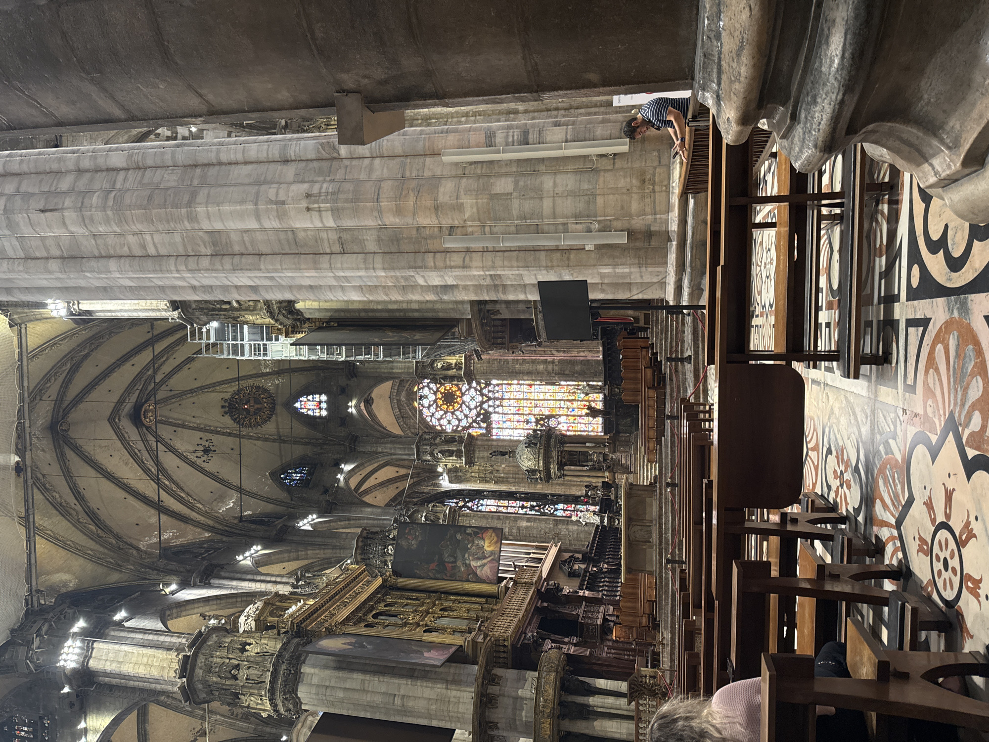 The image depicts the interior of the Milan Cathedral (Duomo di Milano) in Milan, Italy. The vast nave of the cathedral is visible, characterized by soaring vaulted ceilings, large marble columns, and intricate architectural details. Sunlight streams through stained-glass windows, illuminating the ornate floor with decorative patterns. Rows of dark wooden pews are arranged throughout the space, some occupied by visitors. In the foreground, to the left, a person with blonde hair is seated on a pew, facing away from the camera. To the right, near a large marble column, a person wearing a striped t-shirt is looking at their phone while leaning on a railing. The cathedral's grandeur and scale are evident, with scaffolding visible in the background, suggesting ongoing restoration or maintenance work. The overall atmosphere is one of reverence and awe, with a blend of historical architecture and the presence of modern visitors.