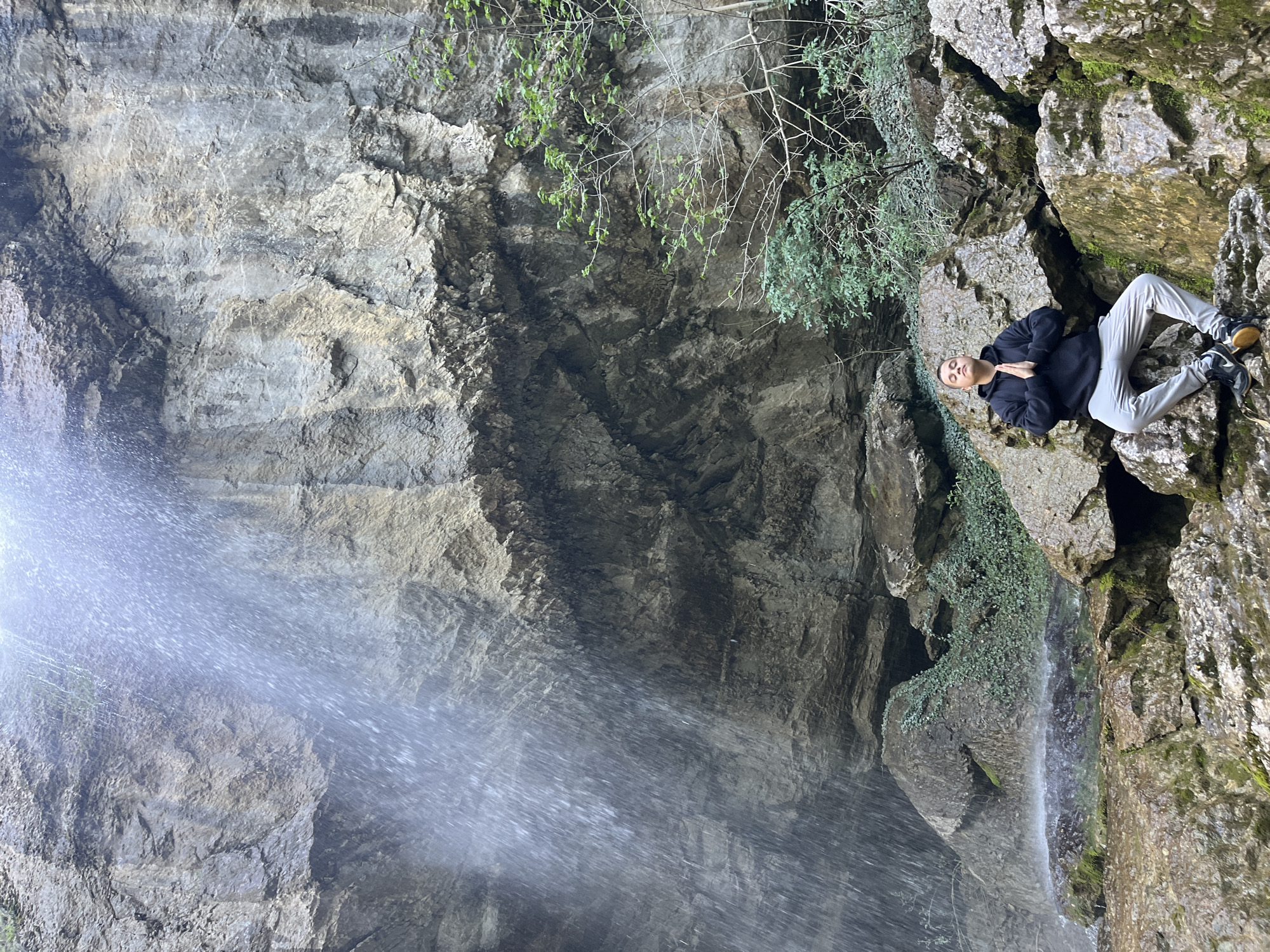 The image shows a man seated on a rocky ledge near a waterfall. The waterfall is partially visible on the left, with water cascading down a steep rock face. The man is wearing a dark hoodie and light-colored pants, and he appears to be in a meditative pose. The surrounding environment consists of moss-covered rocks, foliage, and parts of the cliff wall that form the cave. The location is Veselinovo, Bulgaria.