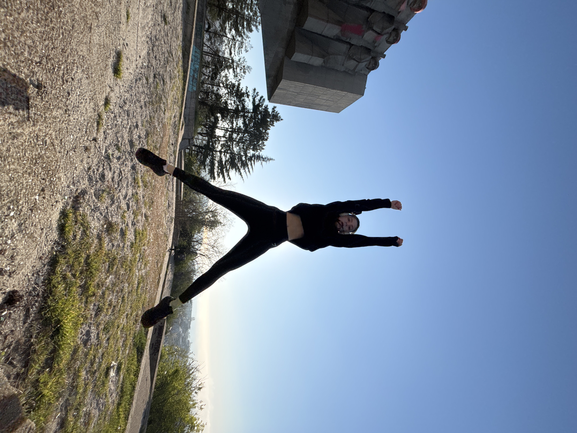 The image shows a person in black clothing with arms outstretched and legs spread, creating a star-like pose against a bright blue sky. The ground, covered in grass and concrete, is visible on the left side of the frame. In the upper portion of the image, there are trees and a large concrete structure. The scene appears to be taken outdoors, possibly in a park or public space. The specific location within the city of Unknown, Unknown is not discernible.