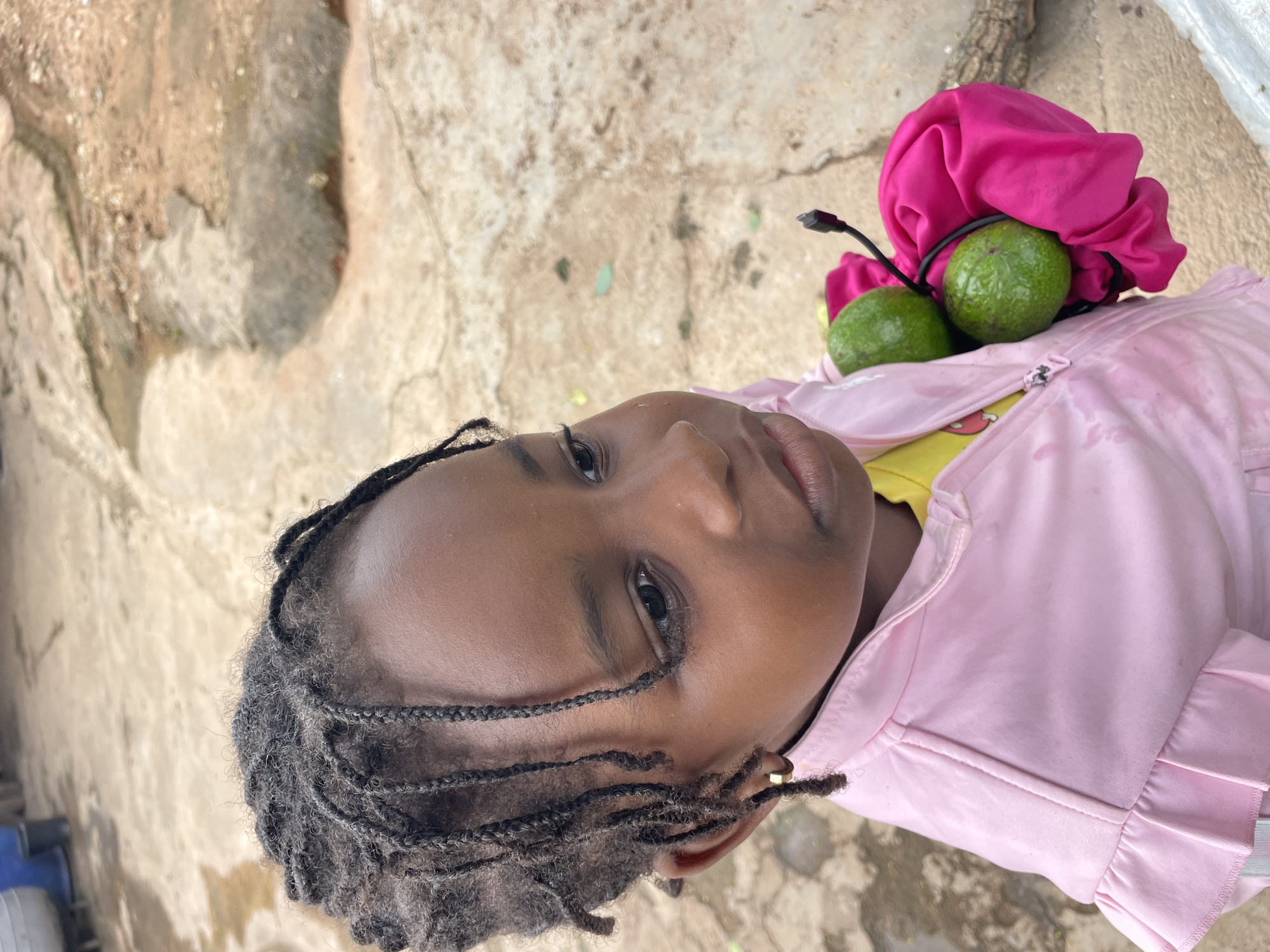 A young Black girl with braided hair and a gold hoop earring in her left ear is holding two green avocados wrapped in a bright pink cloth. She is wearing a light pink jacket zipped up over a yellow shirt. The background appears to be an outdoor setting with cracked concrete ground and some debris, suggesting a utilitarian or informal area. The scene is likely in a residential or open space within the city of Jos, Nigeria, given the context.