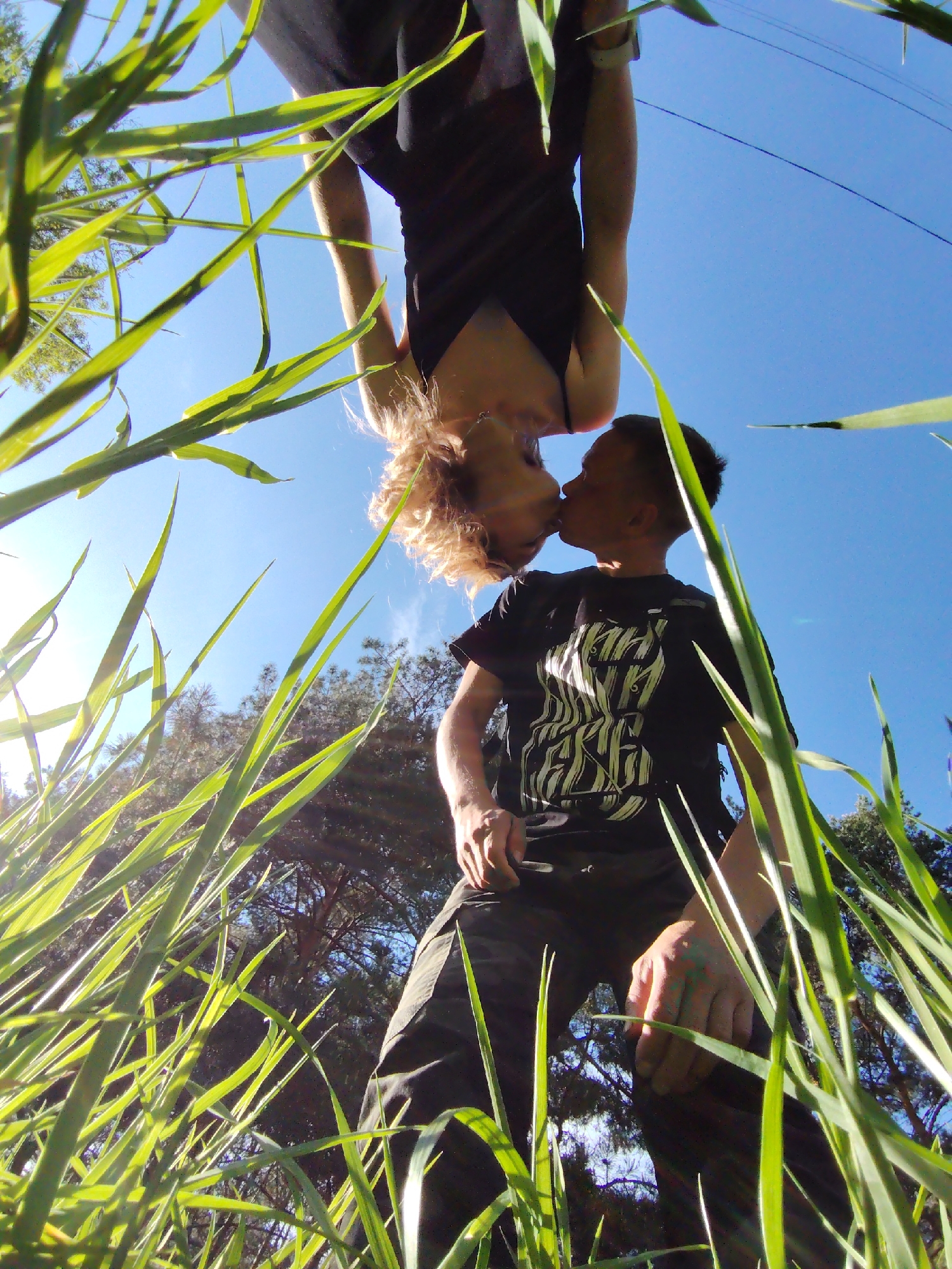A worm's-eye view shows a couple kissing, framed by tall blades of grass. The man wears a black t-shirt with a white design and dark pants, while the woman is above him wearing a dark tank top. The sky is blue with the sun visible on the left. Trees are visible in the background. This image could be captured in the city of Chaikovsky, Russia; this is a presumption only.