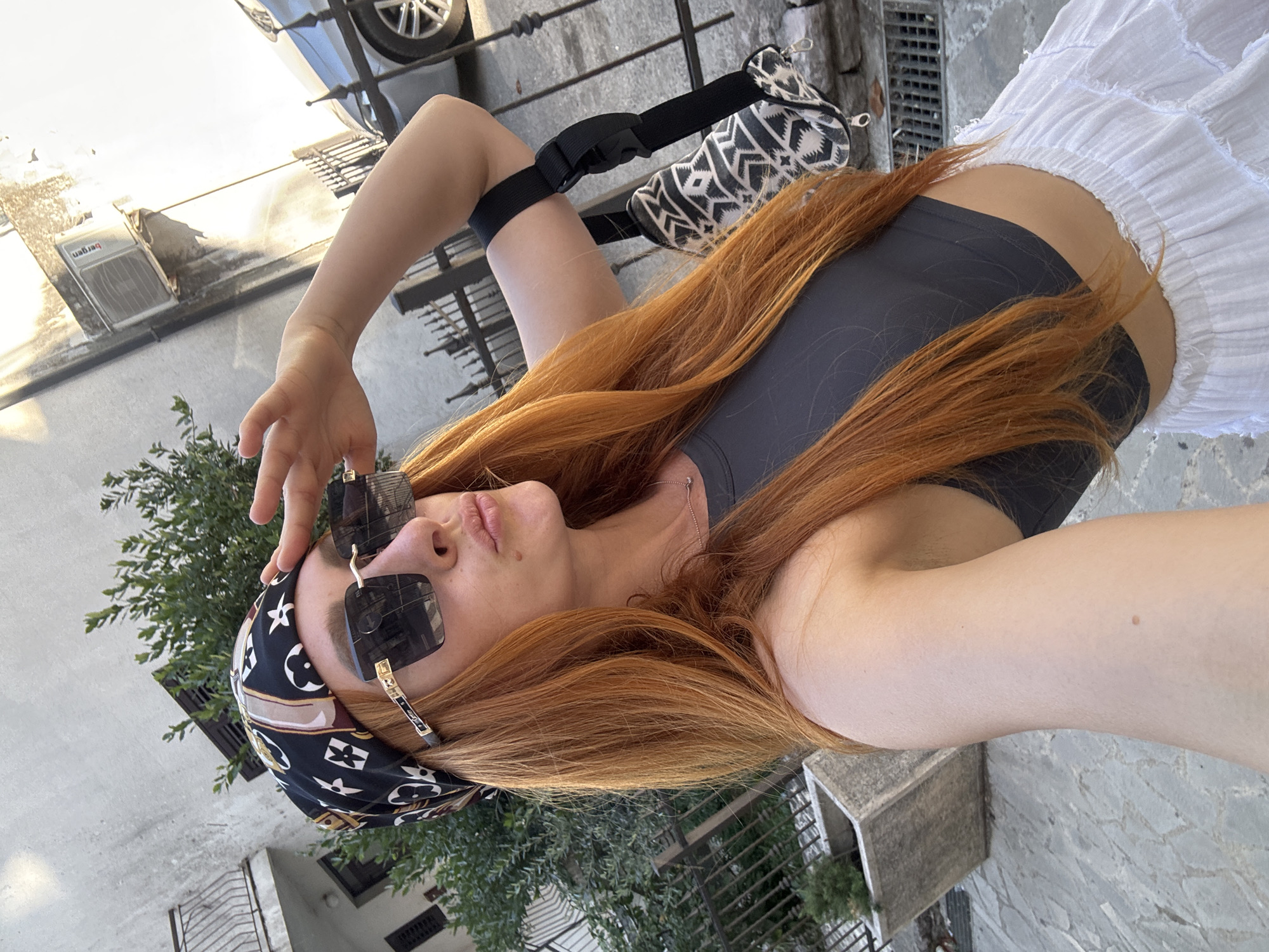 This media file depicts a young woman with long, auburn hair, wearing sunglasses and a headscarf with a designer-inspired print. She is also wearing a dark-colored crop top and a white ruffled skirt. A black utility pouch is attached to her leg with a strap.
The scene appears to be outdoors, possibly on a street or walkway, with a building wall and some greenery in the background. The lighting suggests it is daytime, and the weather seems to be sunny. The woman is looking upwards and appears to be posing for a photo, with her hand touching her forehead. The overall mood is casual and summery.
The headscarf has a pattern that resembles the Louis Vuitton monogram. There are no other discernible text elements in the image. The location context is Budva, Montenegro.
