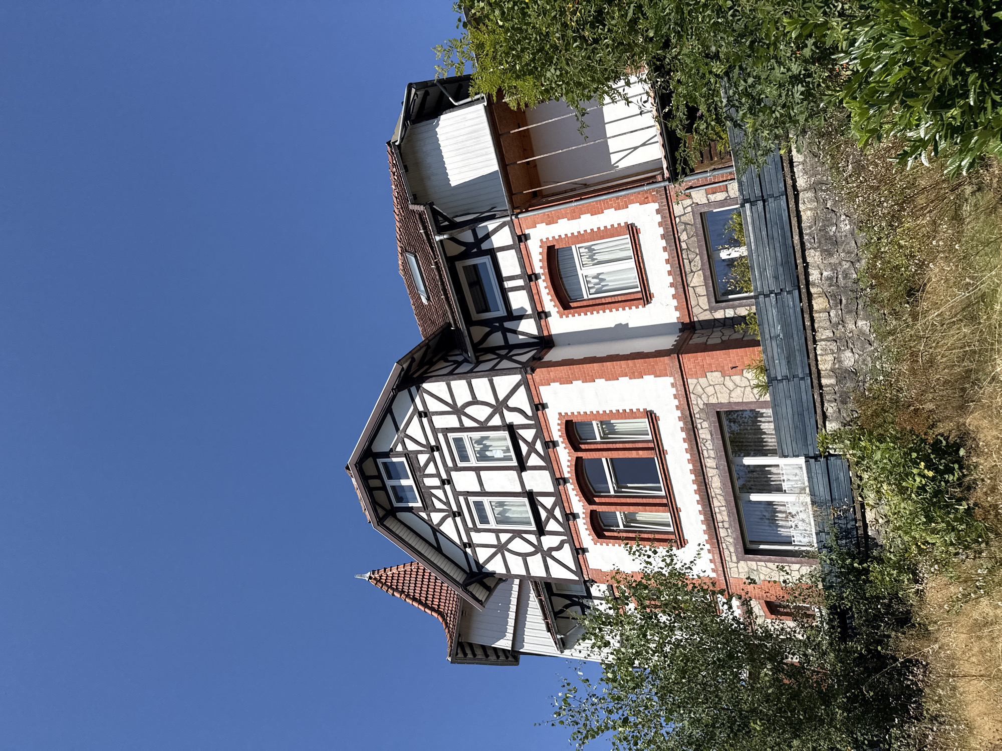 An eye-level, medium shot captures a multi-story building in Bad Sachsa, Germany, against a clear, vibrant blue sky. The building, characterized by its distinctive half-timbered facade with white stucco and dark brown beams, features red brick accents on its lower levels and around the windows. The roof is tiled, with some sections showing signs of age. The architecture is traditional, with several windows on each floor, some adorned with white curtains. A conical red-tiled turret rises from the left side of the building, adding to its unique aesthetic. The structure is partially obscured by lush green trees and vegetation on the right and left, as well as in the foreground. A stone retaining wall with a dark grey wooden fence runs along the front of the property, separating it from the grassy, dry vegetation that covers the sloping ground. The scene appears tranquil and well-maintained, suggesting a residential property or a guesthouse. There are no people visible in the image.