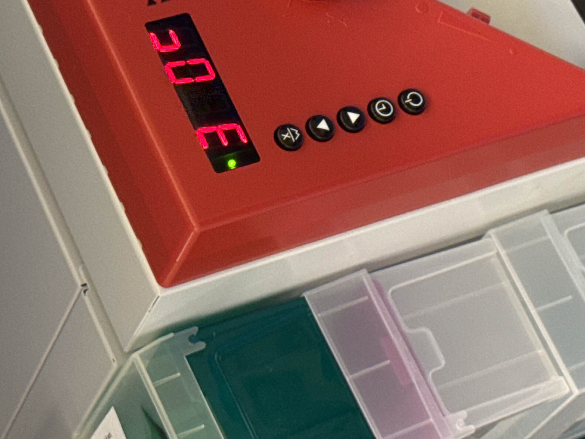 The image presents a close-up, angled view of a red electronic device with a black control panel. The device features a red digital display, which clearly spells out "CODE" using seven-segment characters, and a small, illuminated green LED light directly beneath it, indicating power or active status.
To the right of the display, there are five black circular control buttons with white icons. From left to right, these icons depict: a symbol resembling programming tags (e.g., `< / >`), a left-pointing arrow, a right-pointing arrow, and two subsequent buttons with power or standby symbols (a circle with a vertical line).
The red device is situated atop a series of translucent and clear plastic modular storage compartments or drawers. One of these compartments contains a dark green rectangular object. The setting appears to be an indoor environment, likely a laboratory, workshop, or office, given the type of electronic equipment and organized storage. No human subjects are visible, and there are no discernible location cues within the image itself. The device is powered on, suggesting it is either in operation or ready for use.