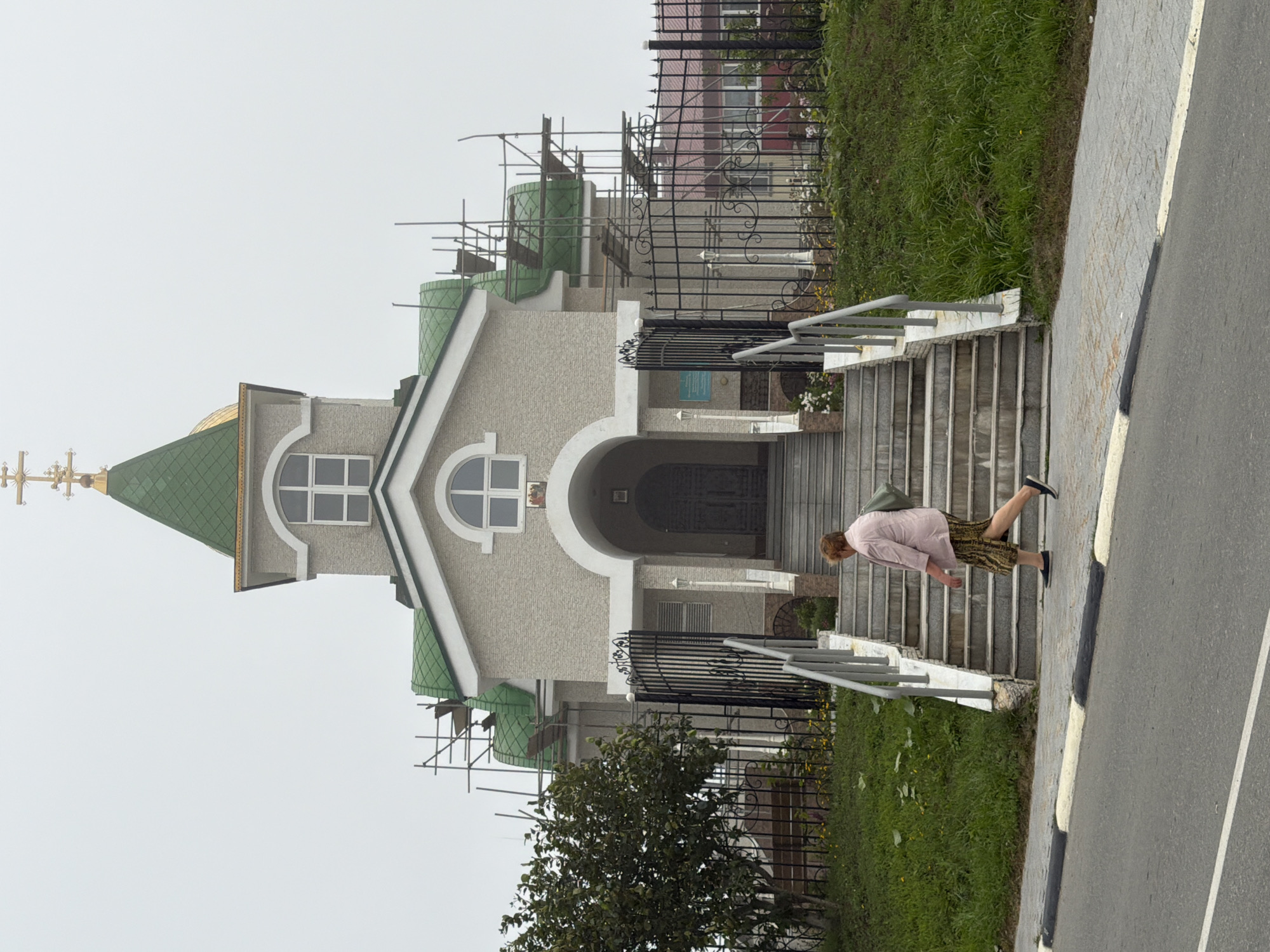 A woman walks down the stairs in front of a church under construction, with scaffolding surrounding the roof. The church has a green roof and a golden cross on top. The woman is wearing a pink jacket and a patterned skirt, and she is carrying a green bag. The church is located in Yuzhno-Kurilsk, Russia. The sky is overcast, and the overall atmosphere is somewhat somber.