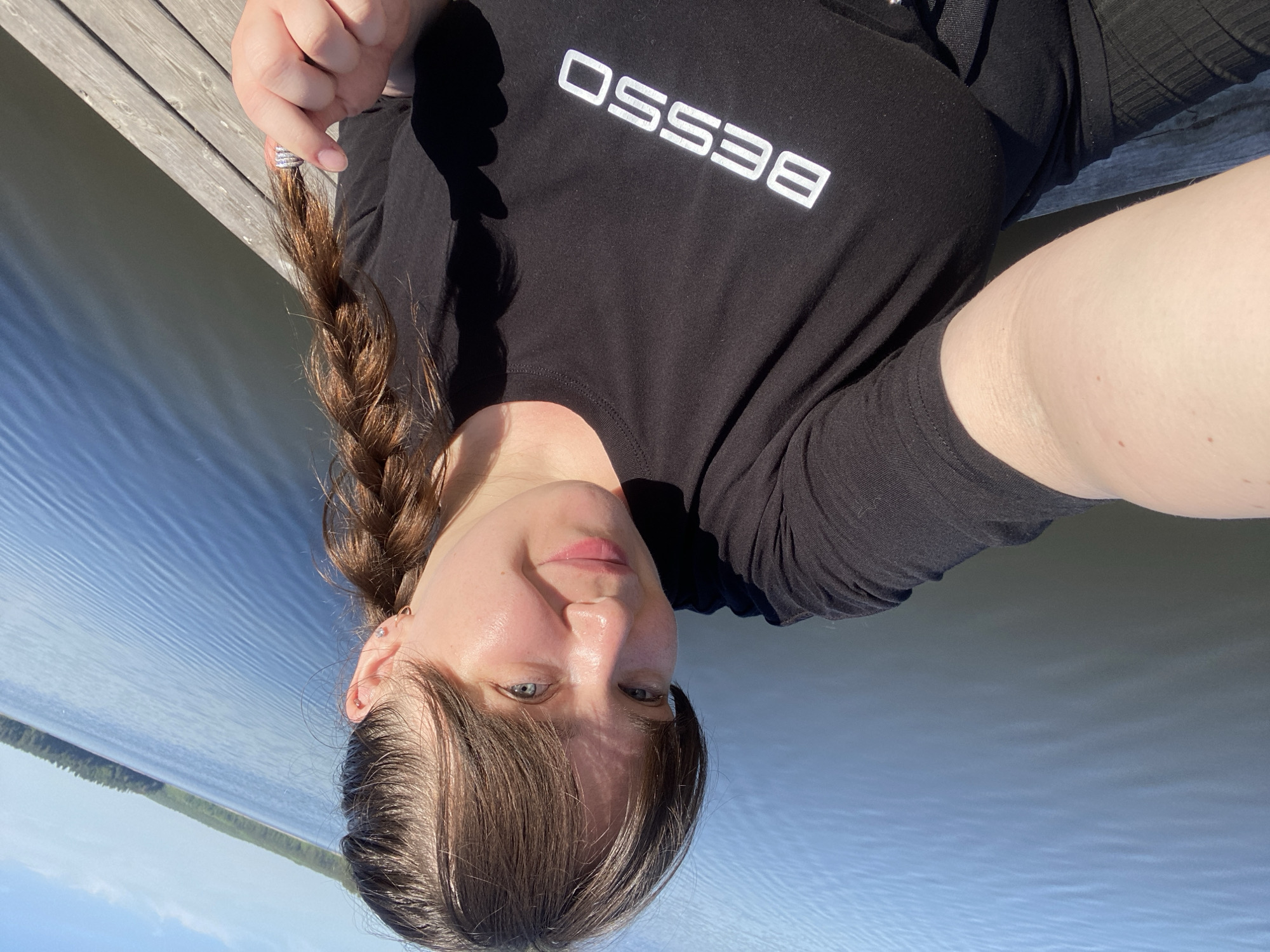 A woman is posing for a selfie, lying on what appears to be a wooden dock. She has brown hair, styled in a braid to the side. She is wearing a black t-shirt with the word "BESSO" printed in white. The background shows a body of water, likely a lake or river, with a treeline visible in the distance. Given the prompt, the location is likely in Votkinsk, Russia.