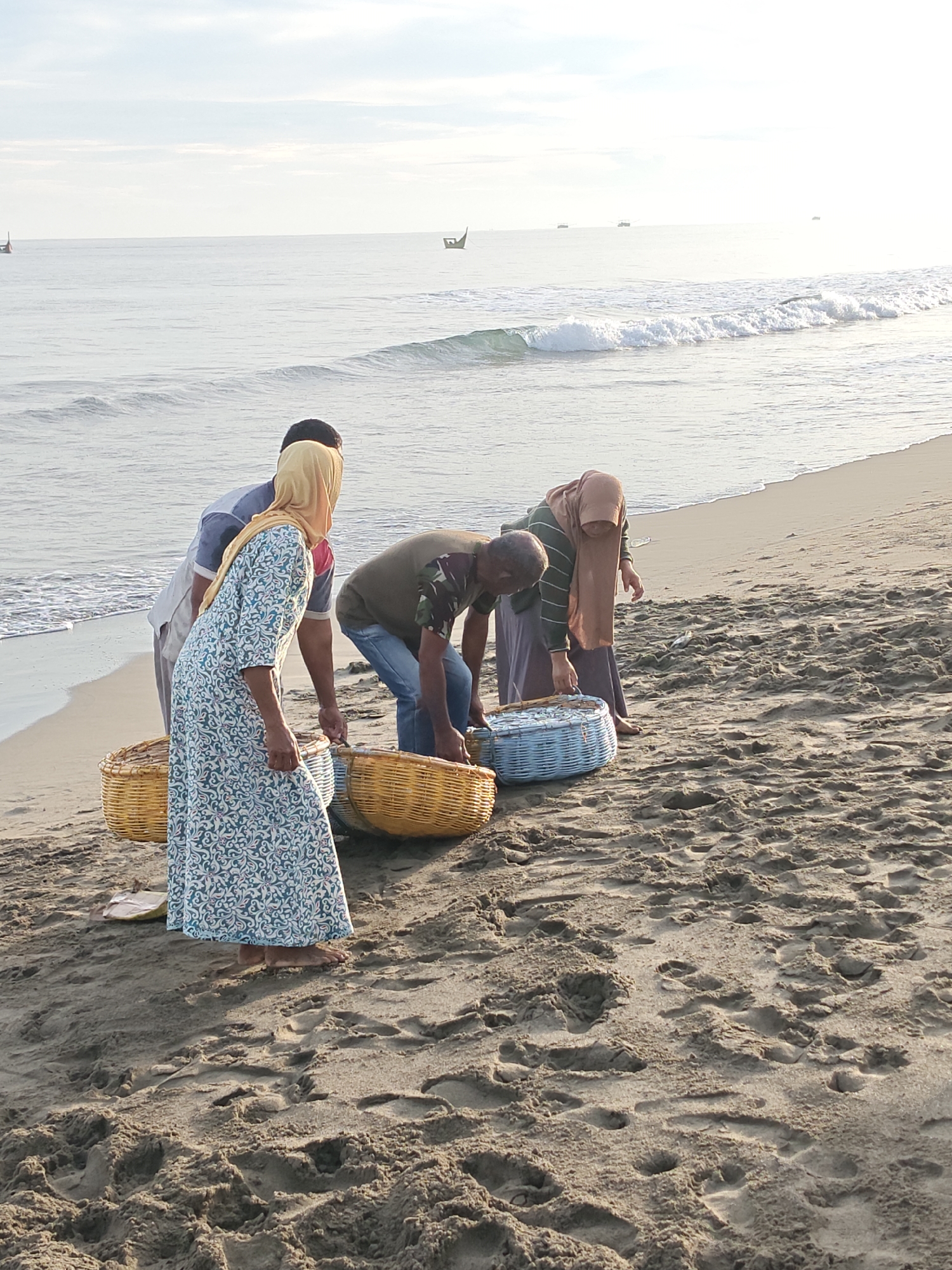 Four individuals are gathered on a sandy beach near Curehtunong, Indonesia, by the ocean. The people, two men and two women, are focused on several woven baskets filled with what appears to be fish. The ocean is calm with gentle waves, and the sky is bright and clear. Two boats are visible in the distance on the water. The scene suggests a fishing activity or the collection of a recent catch.