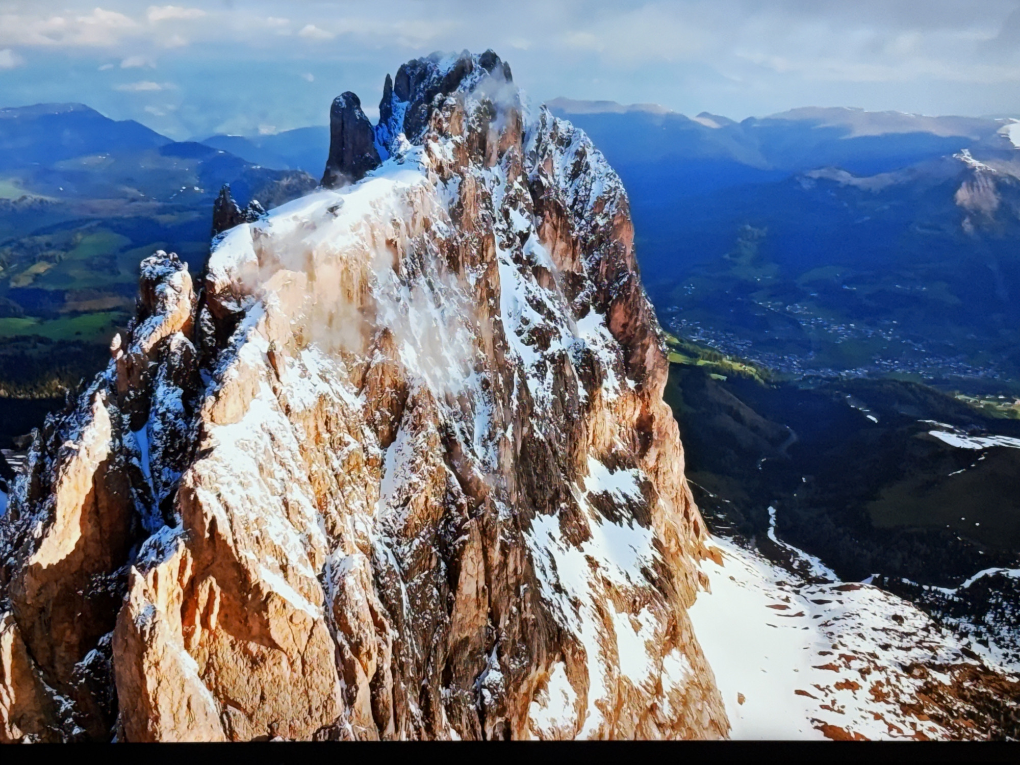 The image depicts a dramatic mountain landscape with snow-capped peaks and rugged rock formations. In the foreground, a colossal, sunlit mountain face dominates the frame, its textures highlighted by the warm light. Patches of snow cling to its slopes, interspersed with dark, sheer rock faces. Wisps of mist or cloud add an ethereal quality to the upper reaches of the mountain.
In the background, a vast expanse of mountains stretches into the distance, their blue hues fading with atmospheric perspective. Below the distant peaks, rolling hills transition into verdant green valleys dotted with a scattering of buildings, suggesting a human settlement or a small town. A winding river or stream is visible in the valley floor. The sky above is a vibrant blue, with scattered white clouds.
This image is an aerial or high-altitude view of a mountainous region. It does not show the city of Burgas, Bulgaria, as Burgas is a coastal city and this landscape is clearly mountainous and inland.