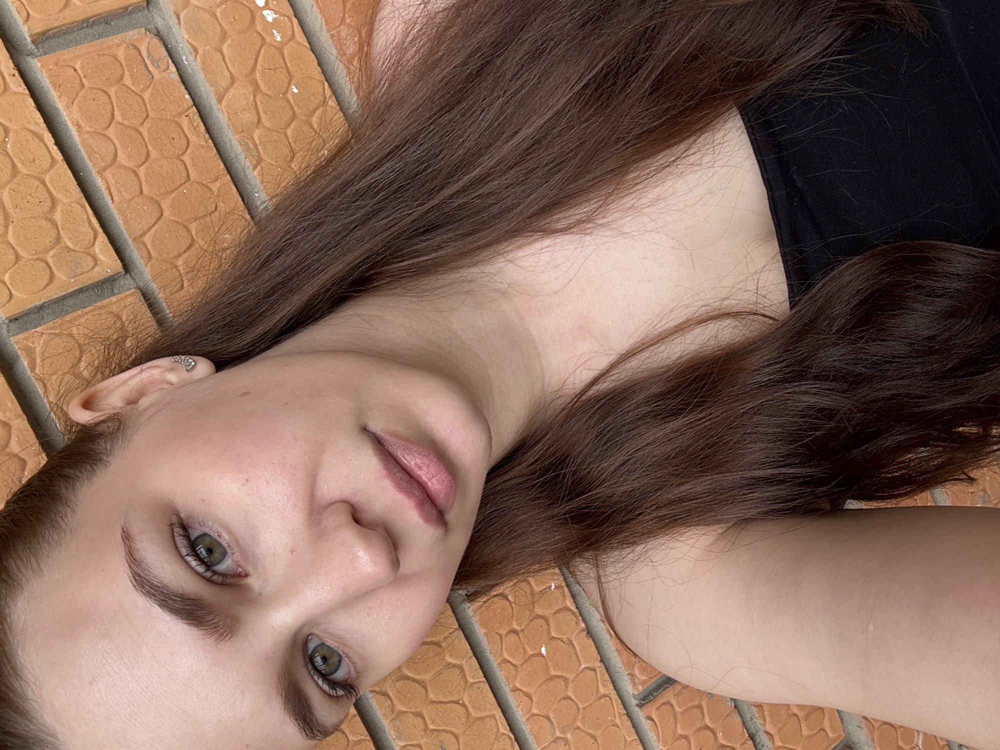 A close-up, angled portrait features a young woman lying on a textured outdoor surface. Her head is positioned in the lower-left, with her body and long, straight, dark brown hair extending diagonally upwards and to the right. The woman has fair skin, green eyes, and naturally defined eyebrows. Her gaze is direct and calm, looking towards the viewer. Her lips are a natural light pink. A small, ornate silver earring is visible on her left earlobe. She wears a black top, a portion of which is visible on her upper body.
The background consists of a reddish-brown brick or tiled floor with a repeating, raised pattern of small, textured shapes within rectangular sections, separated by dark gray grout lines. Some faint white marks, possibly paint or dust, are visible on the surface. The bright, even lighting suggests the photo was taken during the day in an outdoor setting, likely a patio or pathway. The woman appears to be posing for a photograph, conveying a serene and relaxed demeanor. There is no visible text in the image. The location context is Chaikovsky, Russia.