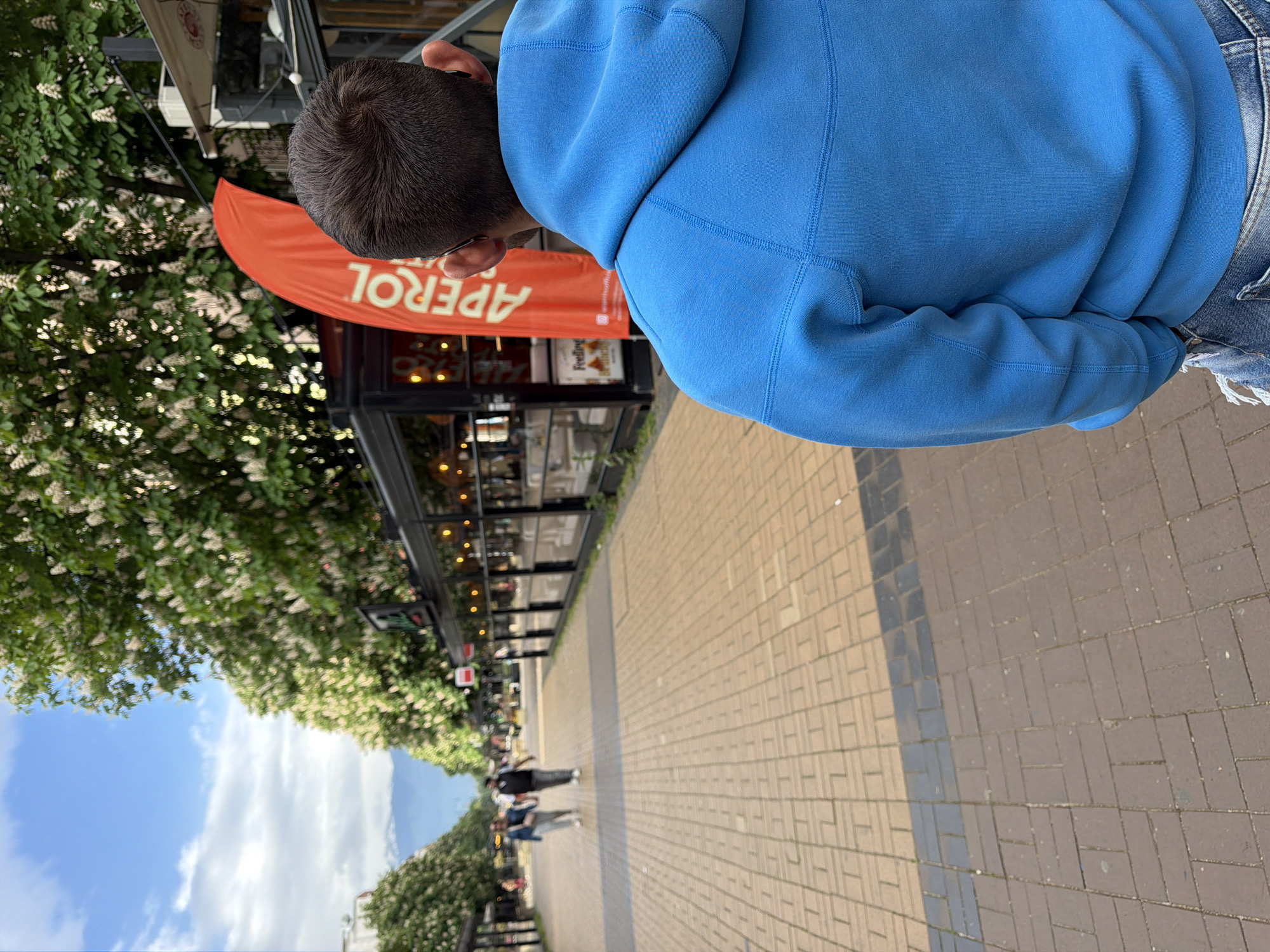 A slightly high-angle shot shows a person wearing a blue hoodie and distressed denim shorts standing on a sidewalk in Sofia, Bulgaria. The person is facing toward an outdoor cafe marked by an orange "Aperol" sign. Along the sidewalk, there are blossoming trees. In the distance, blurred figures of pedestrians are visible. The sidewalk is paved with light-colored brick. Above, there's a view of a blue sky with some clouds.