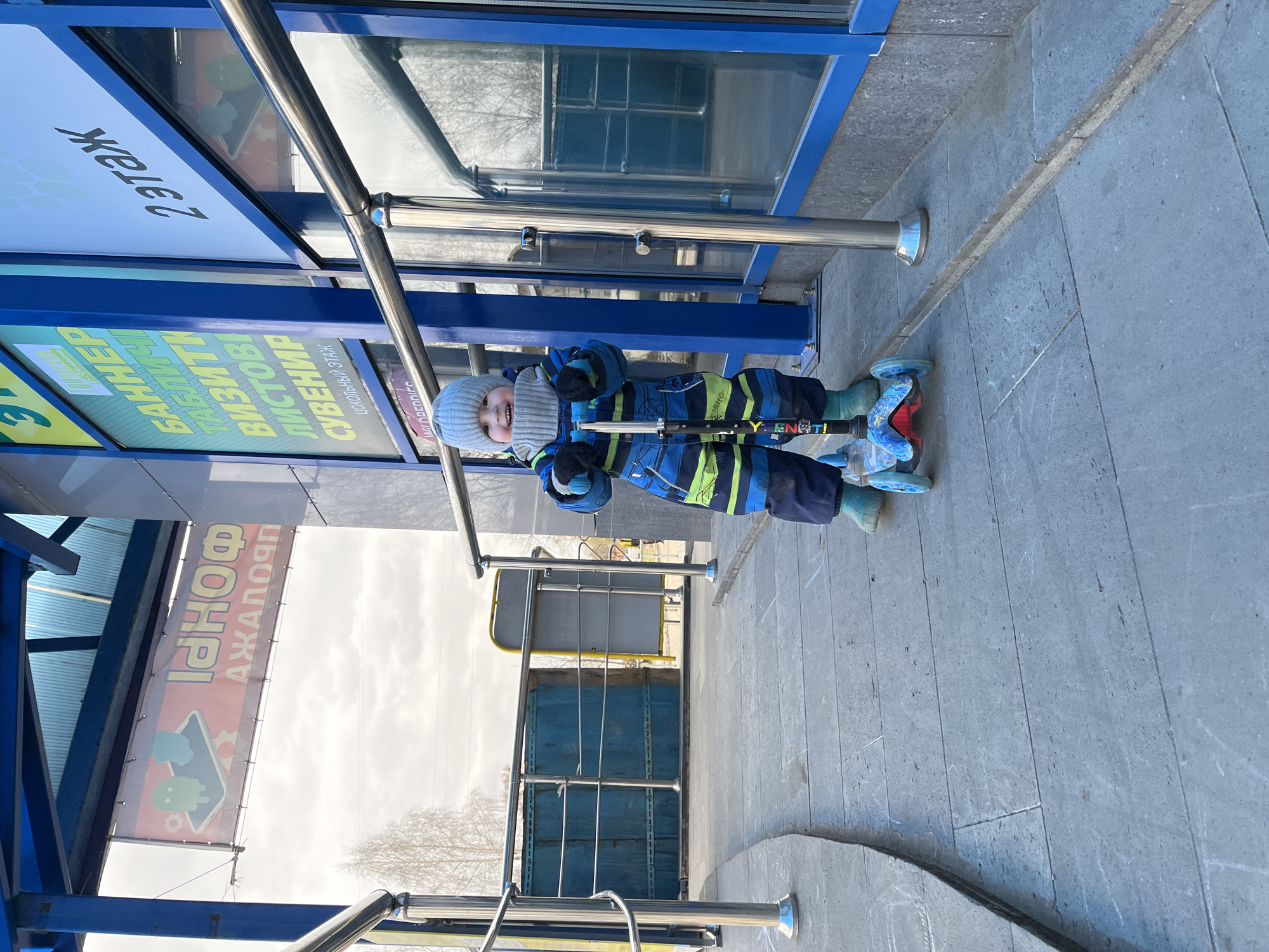 A child stands on a grey tiled sidewalk in front of a building, holding a scooter. The building has blue framing and glass panels, with signs in Russian visible above. The sign on the left reads "Iphonoph Azhapor". Additional signs read "2 floor", "BANNER", "VISITORS", and "SOUVENIR". The child is wearing a blue and green striped jacket, dark pants, and a gray knit hat. The scooter is blue and black. The photo appears to be taken outdoors on a sunny day, with trees visible in the background. The location is in Chaikovsky, Russia, evidenced by the Russian signage.