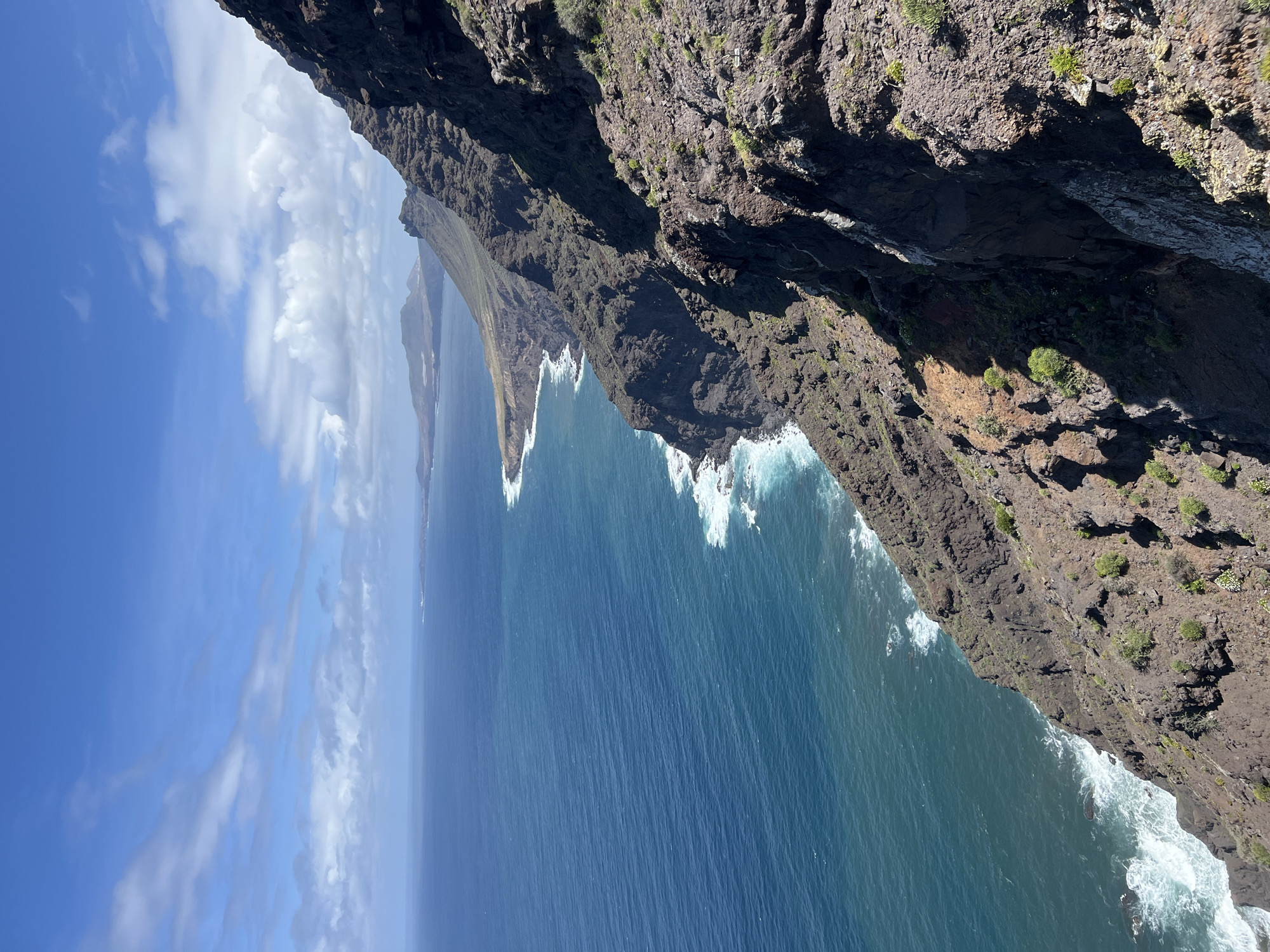 A high-angle view looking down from a steep cliff towards the Atlantic Ocean, with a rugged coastline extending into the distance. The image is dominated by the dark, rocky cliffs in the foreground and midground, with sparse vegetation clinging to the cliff faces. White waves crash against the base of the cliffs. In the distance, a hazy blue sky with patches of white clouds is visible. The location is Tirma, Spain.
