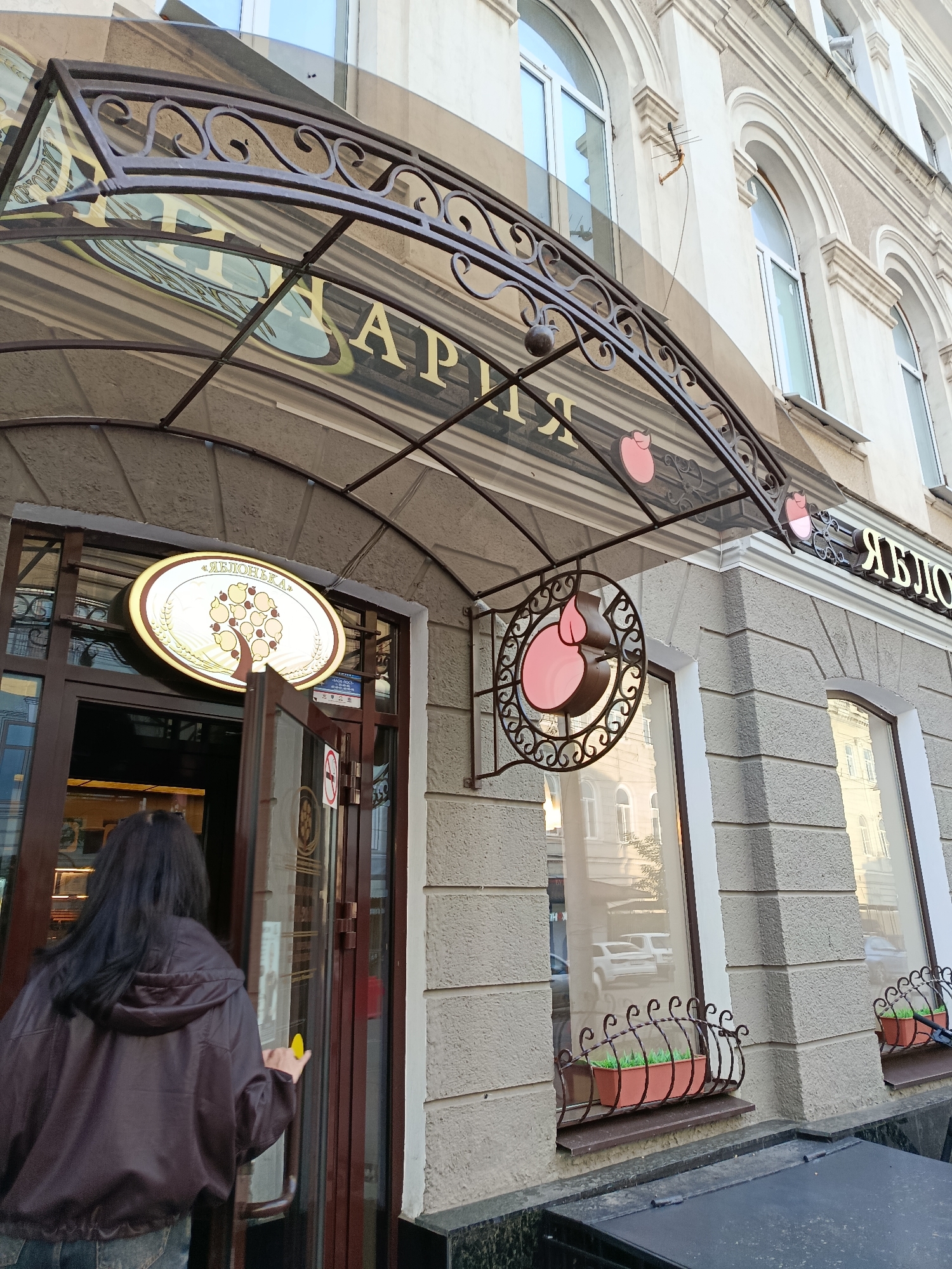 The image shows the facade of a bakery named "Яблонька" (Yablonka) in Saratov, Russia. The bakery is located on the ground floor of a light-colored building with arched windows. Above the entrance, there is an awning with decorative wrought-iron elements and the bakery's name written in gold letters. A round illuminated sign with an apple tree motif is mounted above the entrance door, which is partially open. A woman with long dark hair, wearing a brown hooded jacket and jeans, is entering the bakery. To the right of the entrance, there are windows with flower boxes containing green plants.