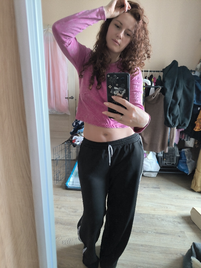 A female individual with curly, reddish-brown hair is depicted in a full-length mirror selfie within an indoor room. She is wearing a pink, long-sleeved, textured crop top that exposes her navel, and black athletic sweatpants with a white drawstring and white stripes along the outer seams. Her left hand is raised, with fingers touching her hair near the temple. Her right hand holds a black smartphone, featuring a stylized graphic design on its case, positioned to capture the image. A silver-colored ring is visible on her right ring finger. The background reveals a somewhat cluttered room with light-colored walls and a light wooden or laminate floor. Visible objects include a metal pet crate, a pile of blue-and-white patterned fabric (possibly laundry), and a clothing rack laden with various garments, including a dark green turtleneck sweater. A light-colored wooden door is partially visible to the far left, behind which a sheer pink fabric element hangs. The scene provides no specific architectural or contextual details to identify a precise location within the city of Saint Petersburg, Russia.